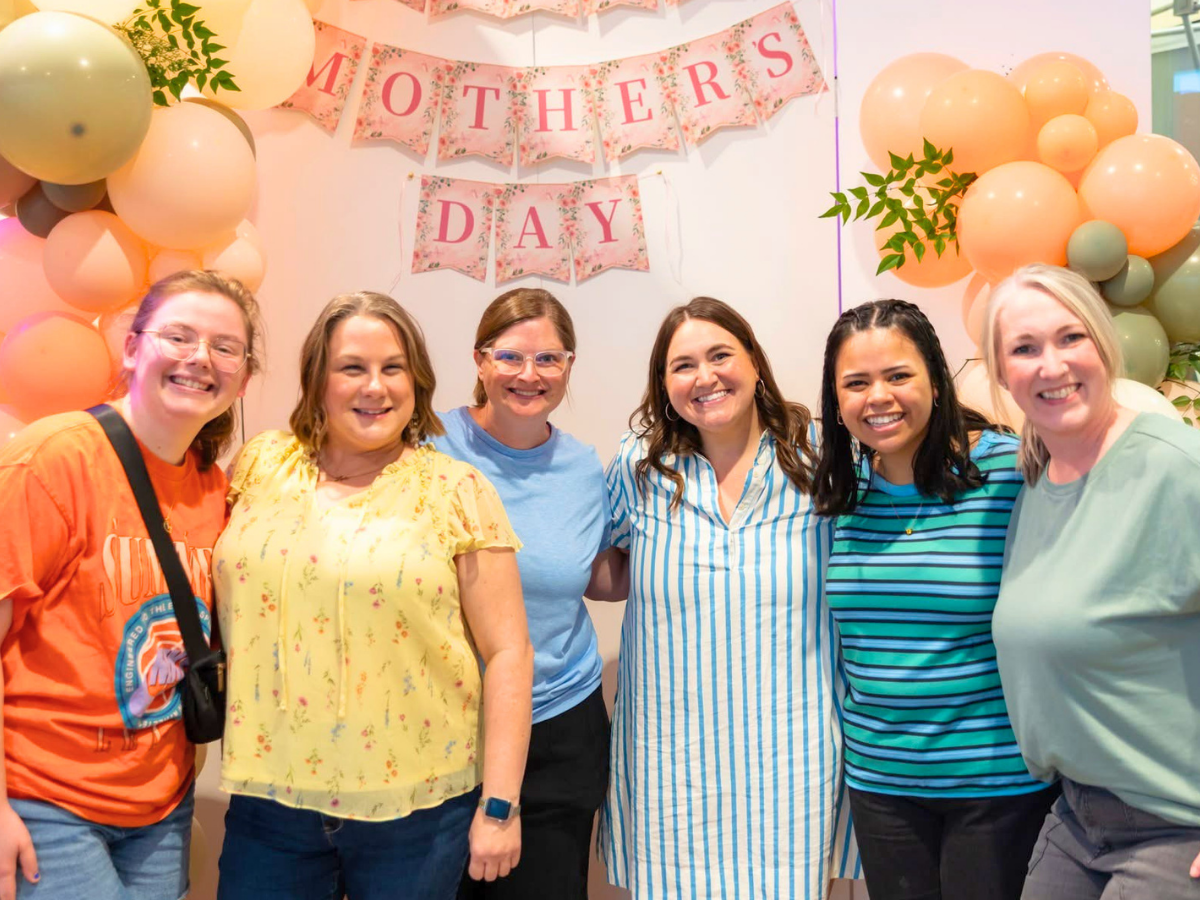 Six women smiling in front of a Mother's Day backdrop with pink banners and pastel balloons in a festive setting.
