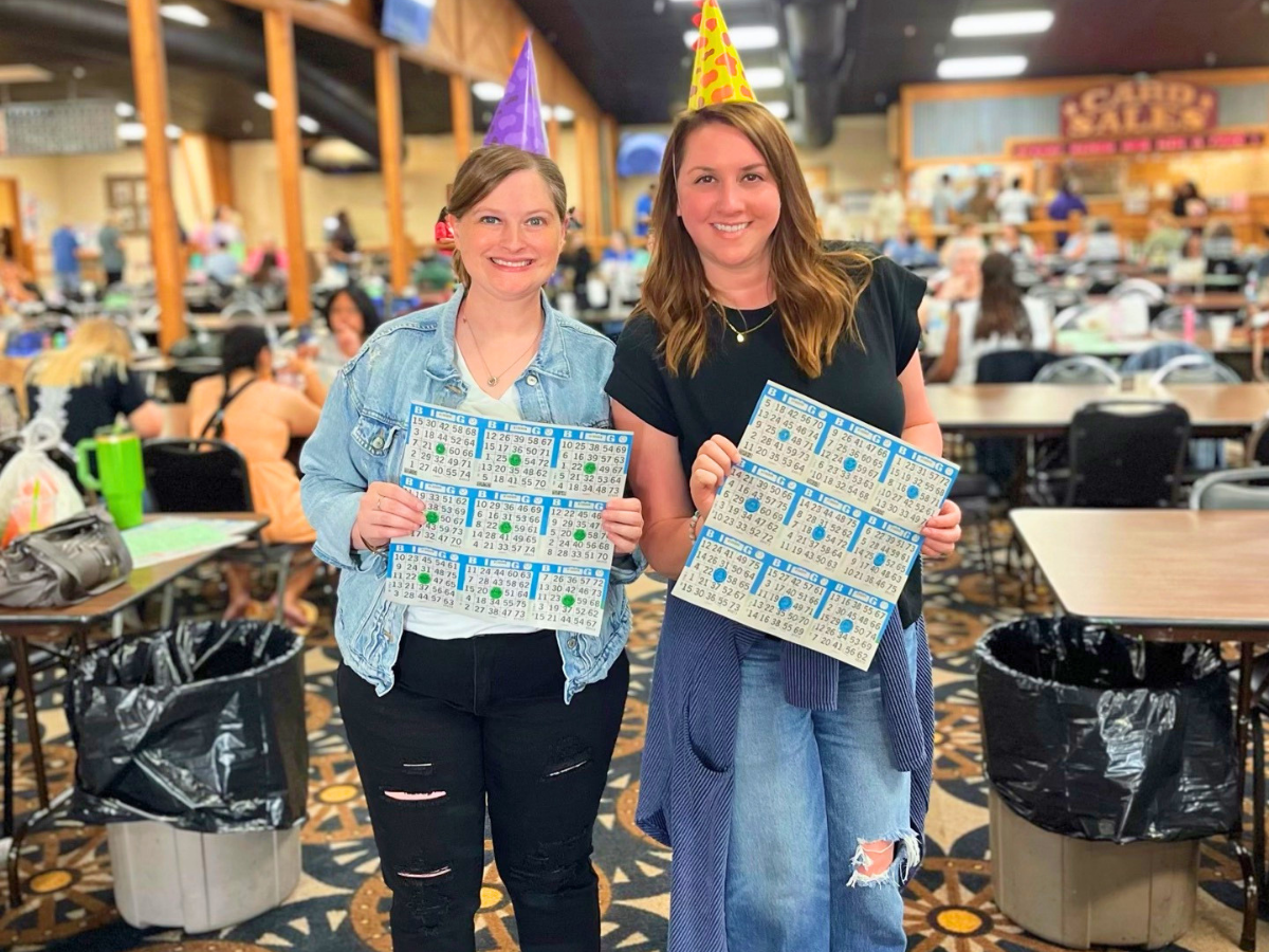 Two women wearing party hats hold up bingo cards and smile in a bustling bingo hall.