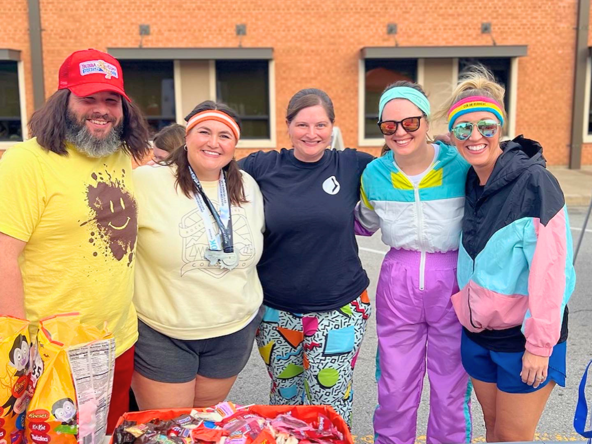 Five adults smiling and posing together outdoors in colorful retro workout outfits, with candy bags in front.