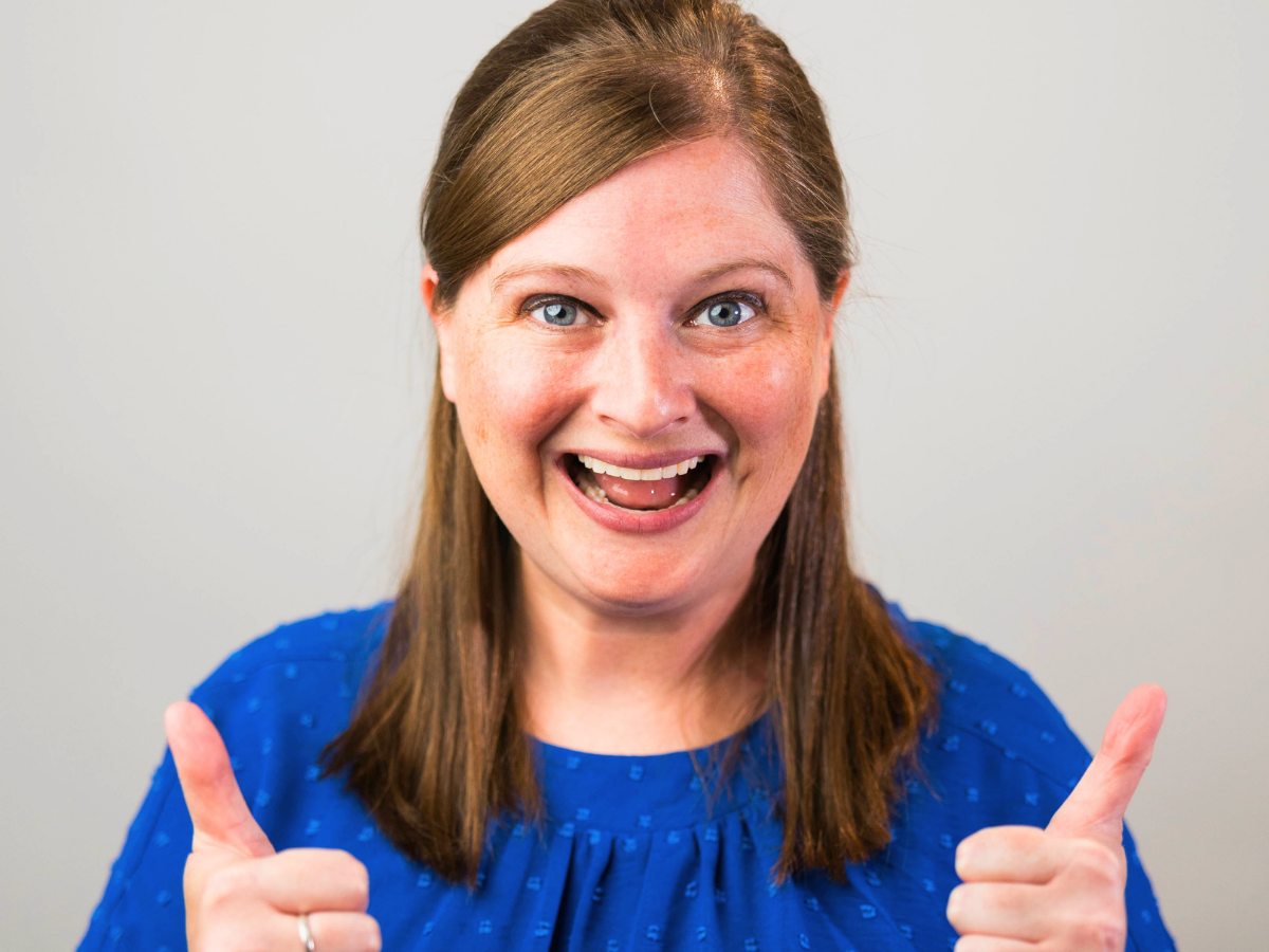 Smiling woman in a blue blouse giving two thumbs up toward the camera.