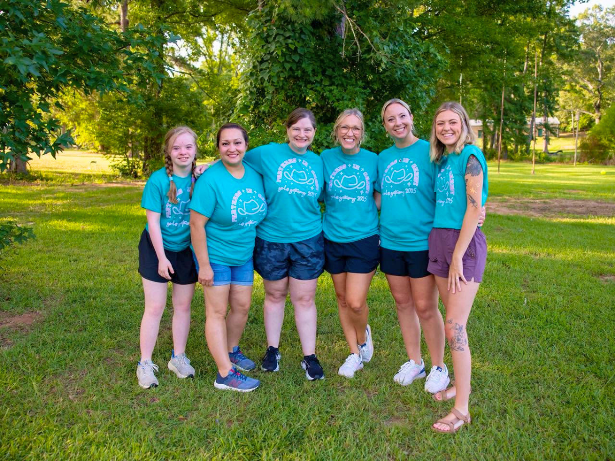 Six women stand together in a sunny park, wearing matching turquoise T‑shirts and shorts, smiling at the camera.