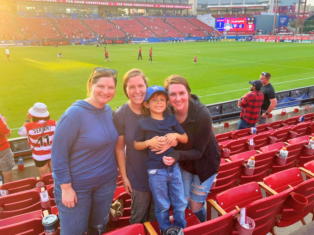 Four smiling people (three women and a young boy) pose together in red stadium seats at a baseball field, with players on the green grass and a scoreboard in the background.