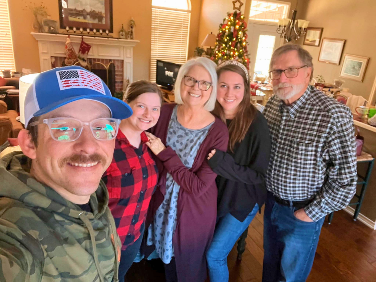 Five adults pose for a smiling group selfie in a cozy living room with a Christmas tree and stockings on the mantelpiece in the background.
