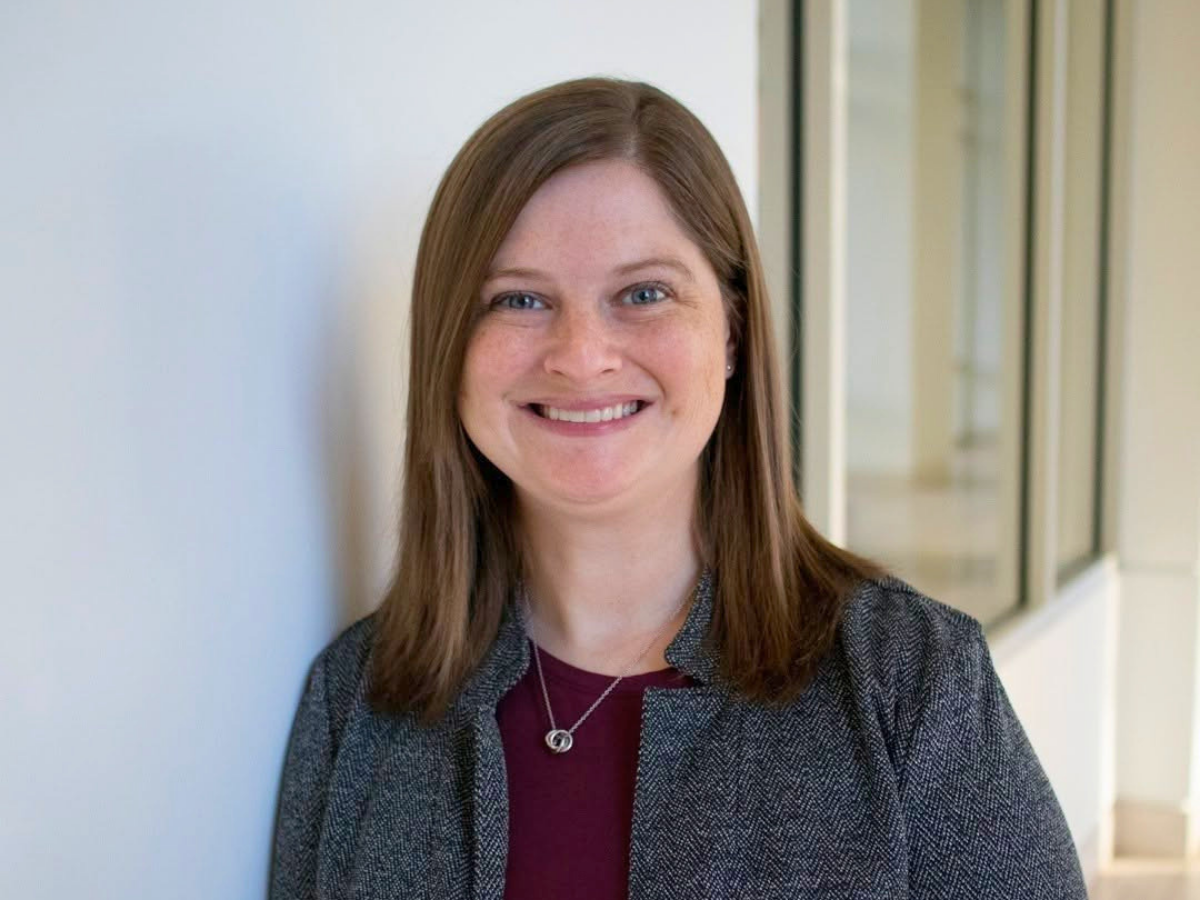 Smiling woman leaning against a light wall in a hallway, wearing a dark blazer and burgundy top.