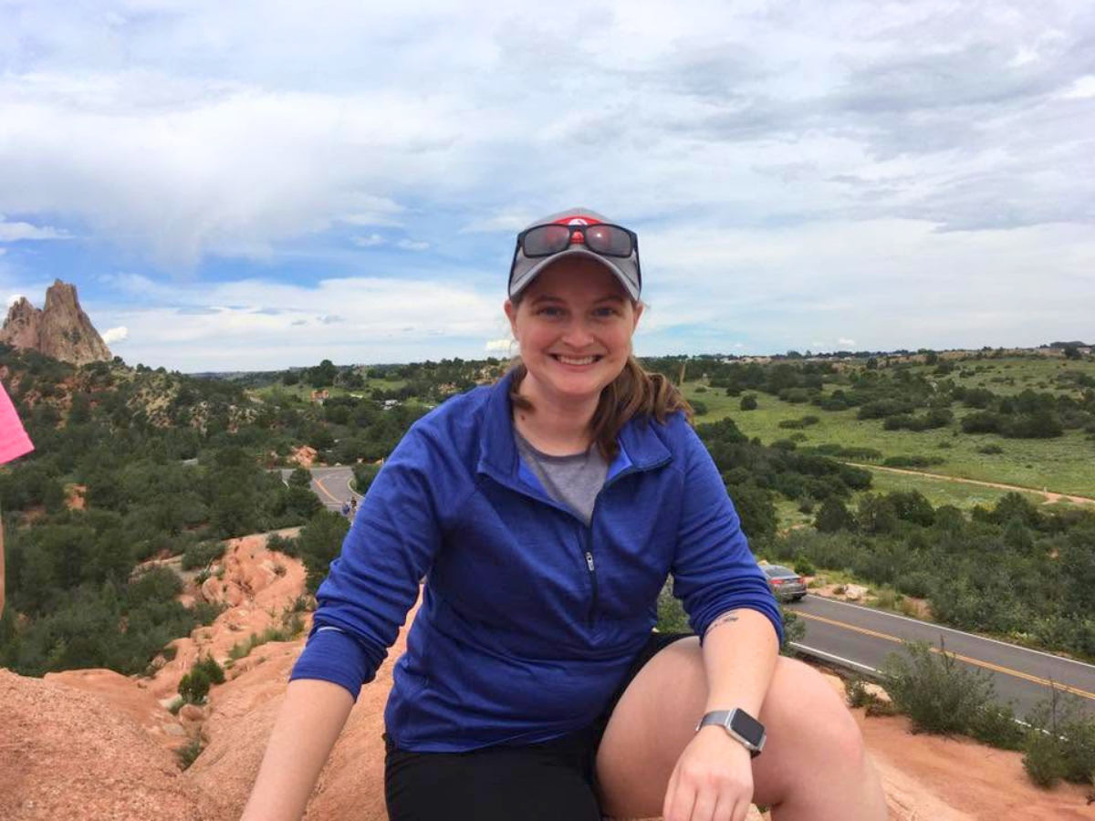 Smiling woman in a blue jacket sits on red rock, with a desert landscape and road in the background under a cloudy sky.