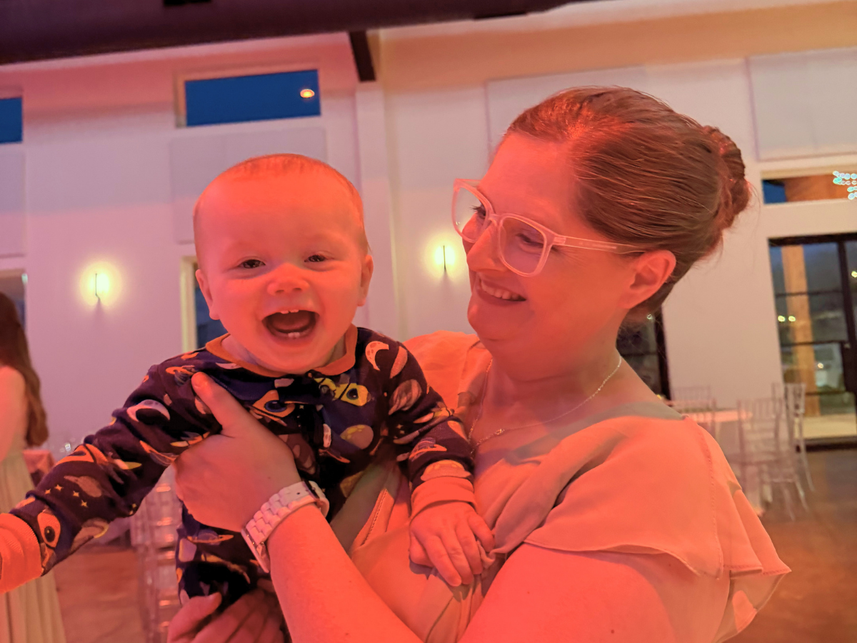 Woman with pink glasses holds a laughing baby at a warmly lit indoor event, both smiling at the camera (reception/party setting).