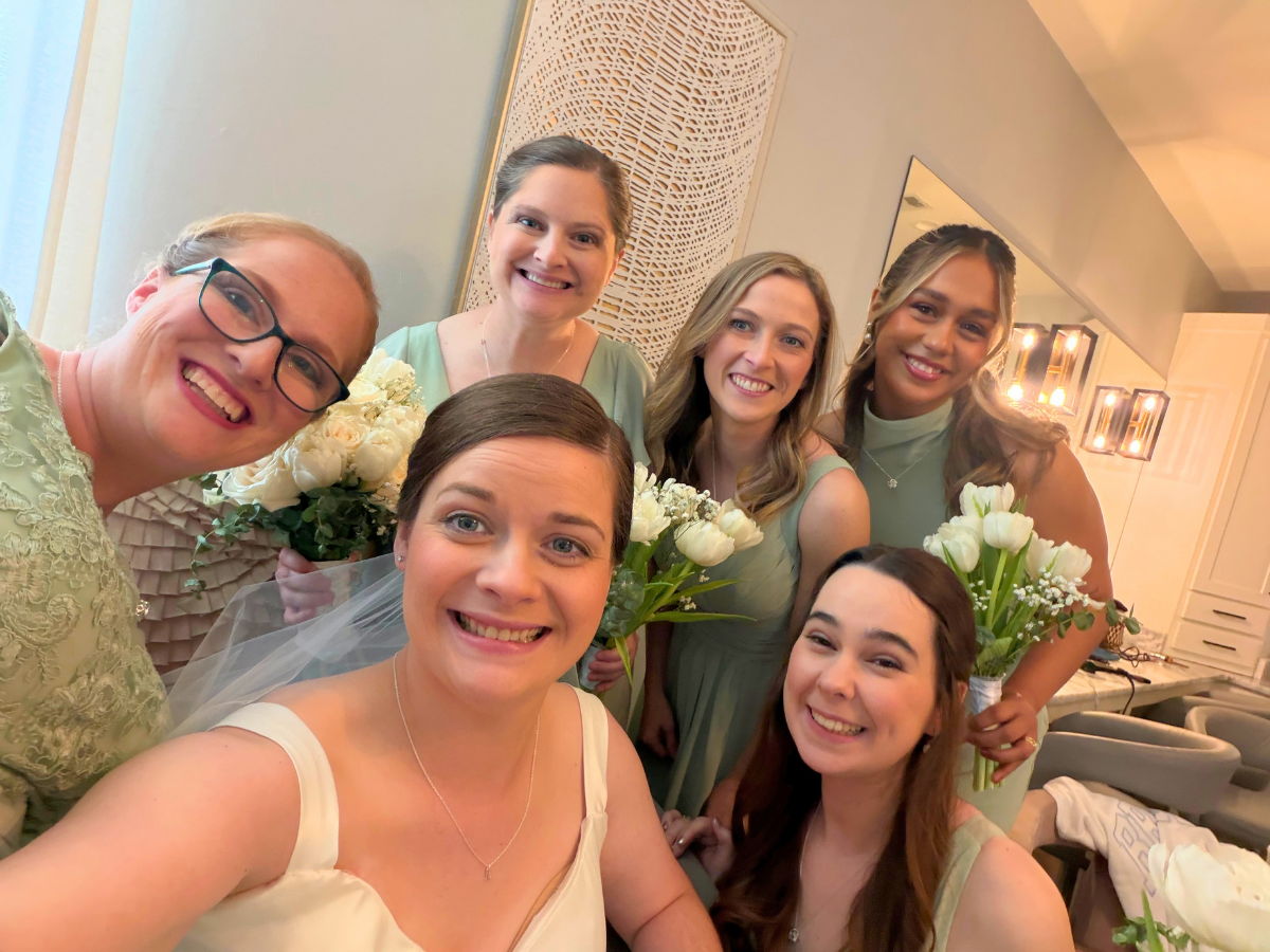 Bride in white dress taking a selfie with six bridesmaids in pastel dresses, each holding white bouquets in a warmly lit room.