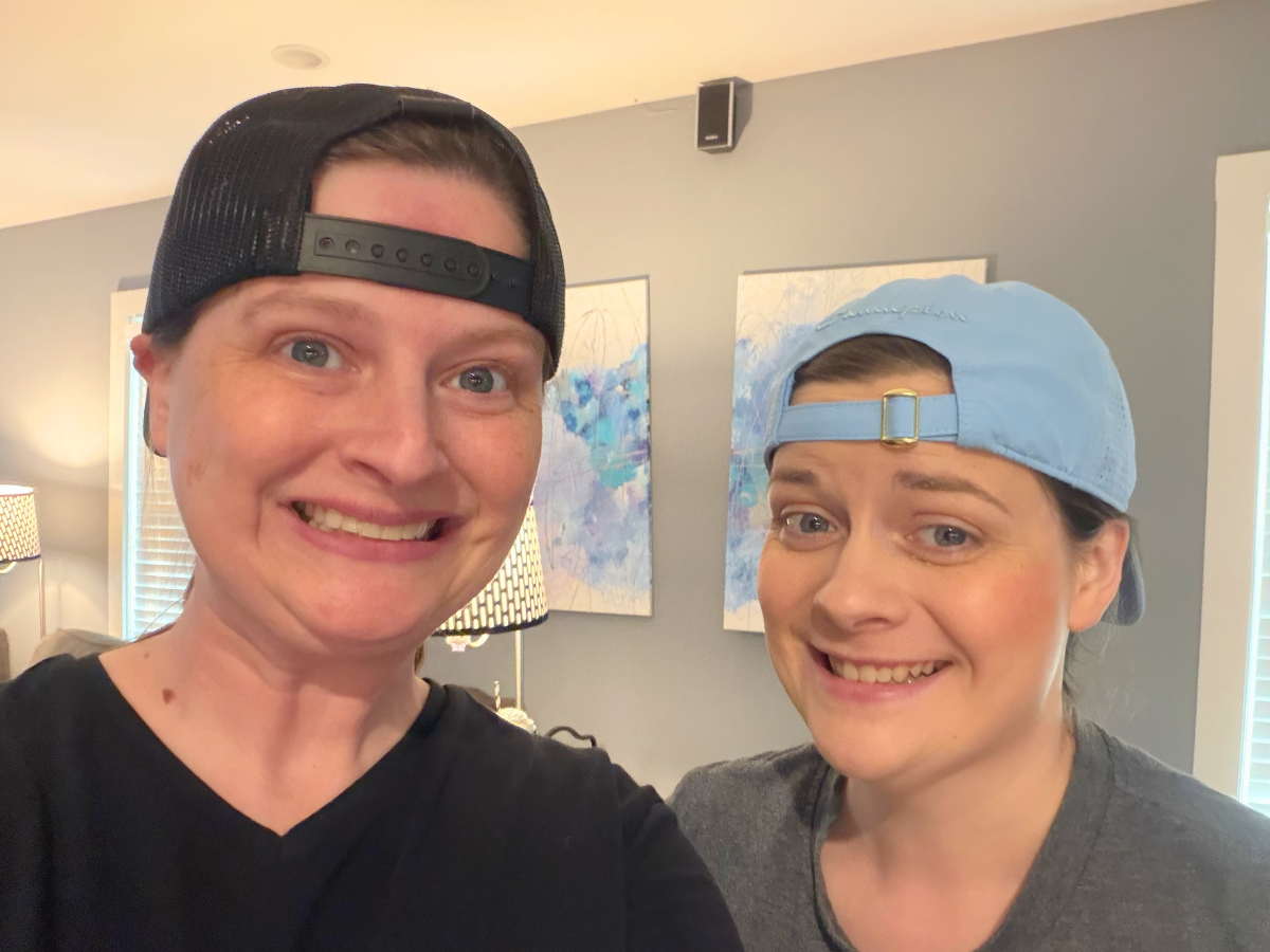 Two people smiling at the camera indoors, wearing backward baseball caps and casual shirts.