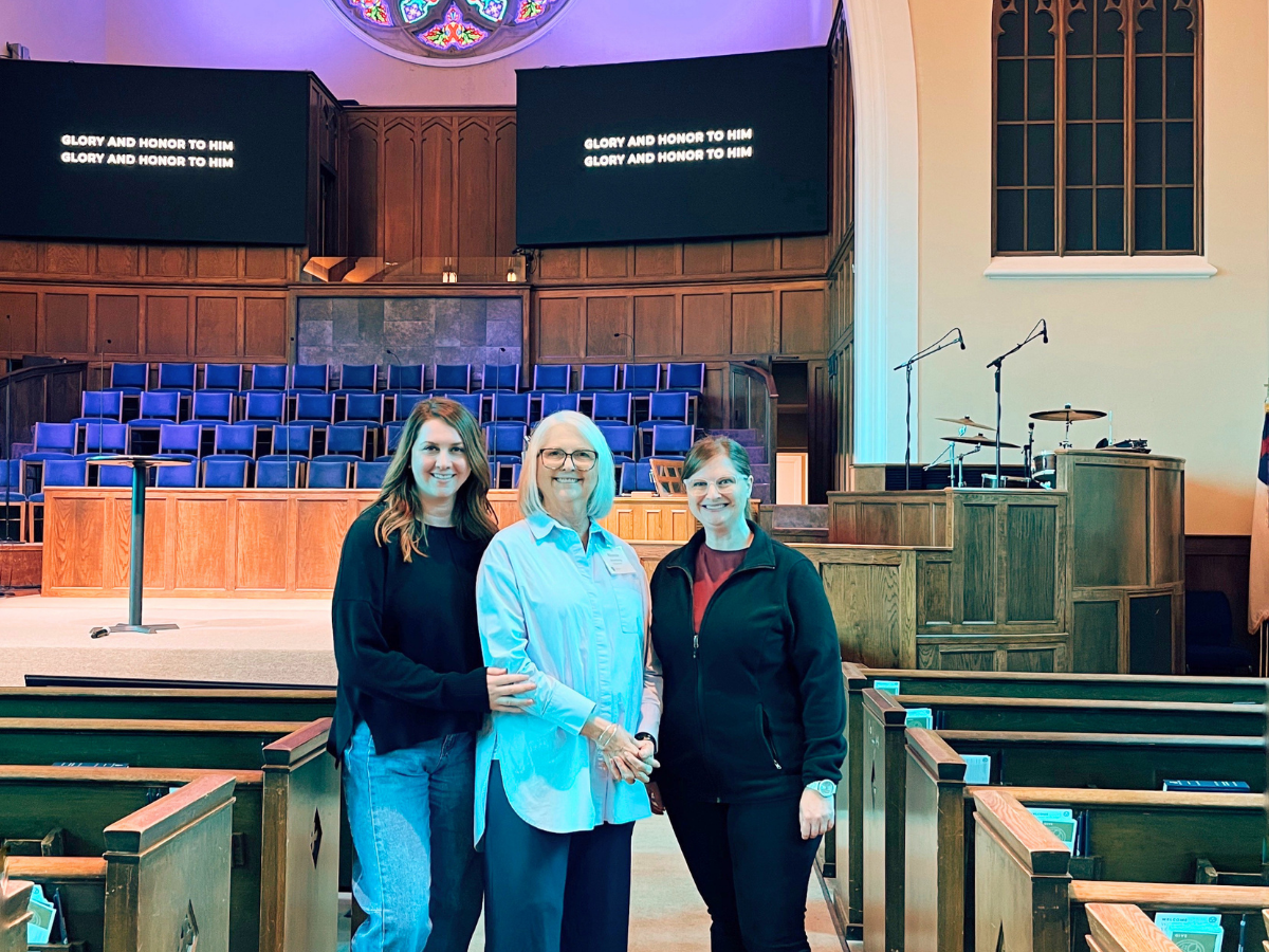 Three women pose together in a church sanctuary with wooden pews and a stage; two screens behind them read “Glory and honor to Him.”