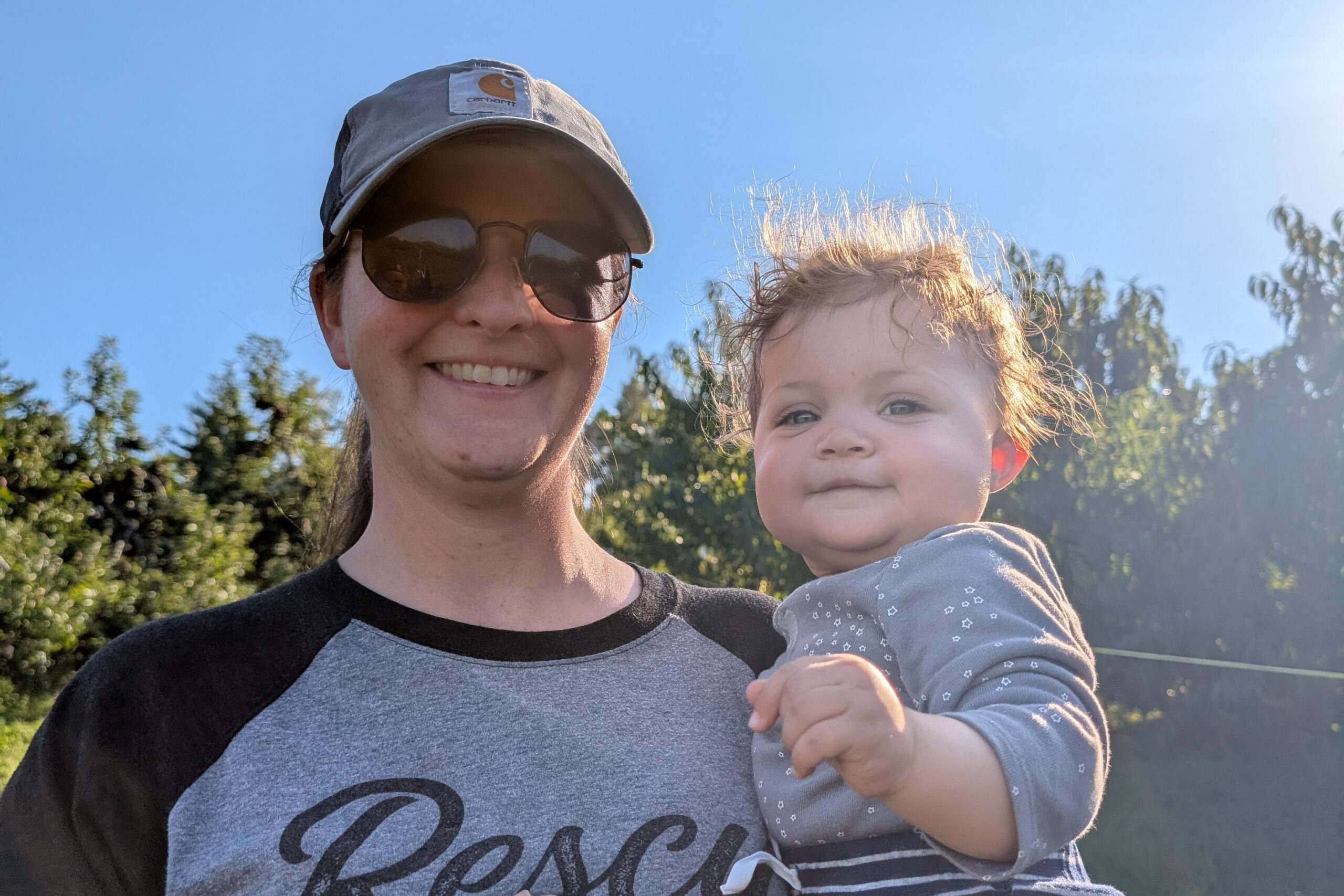 Woman wearing a gray Carhartt cap and sunglasses, smiling while holding a baby outdoors with trees and blue sky in the background.