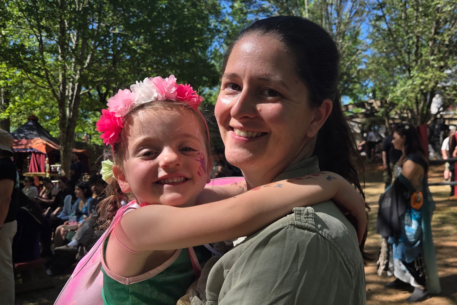 Smiling girl with a pink flower crown hugs a smiling woman at an outdoor festival in the park.