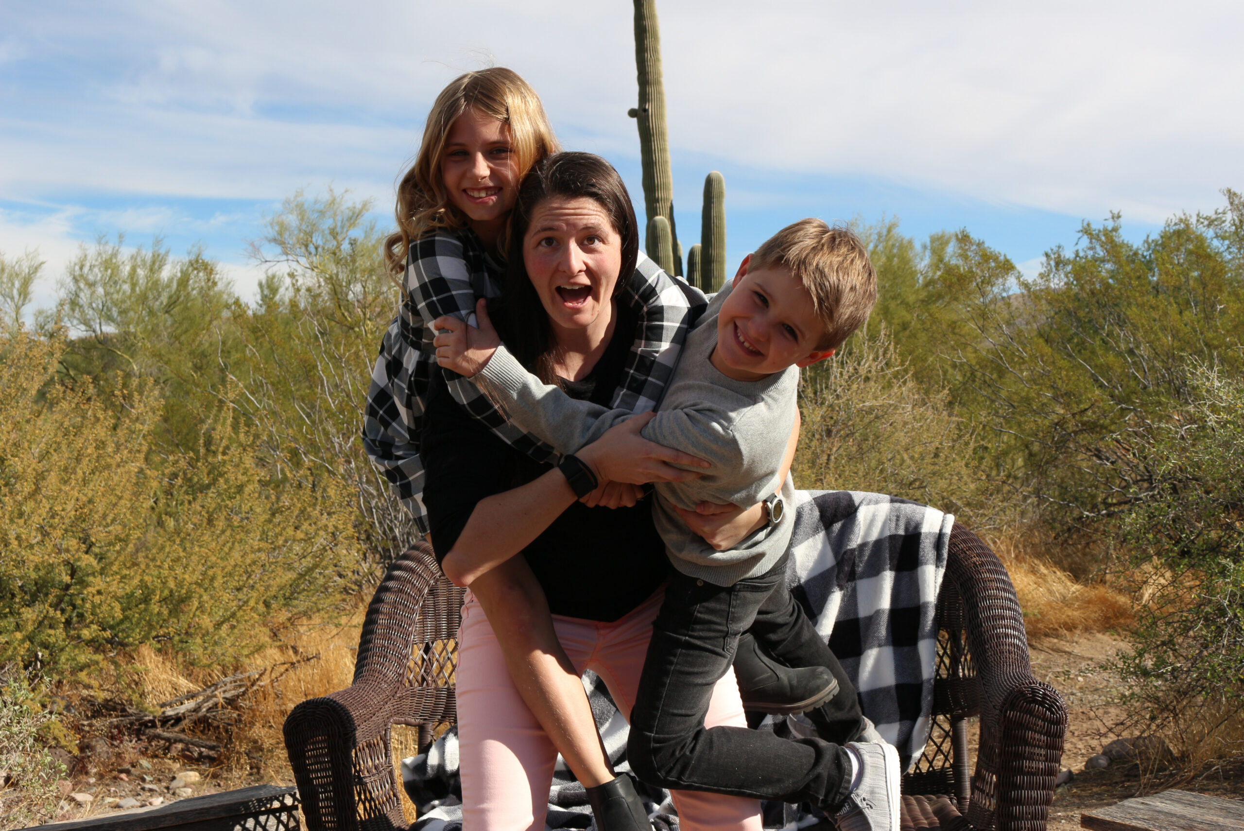 Woman carries two smiling kids on a wicker bench in a desert landscape with tall cacti and dry bushes behind them.
