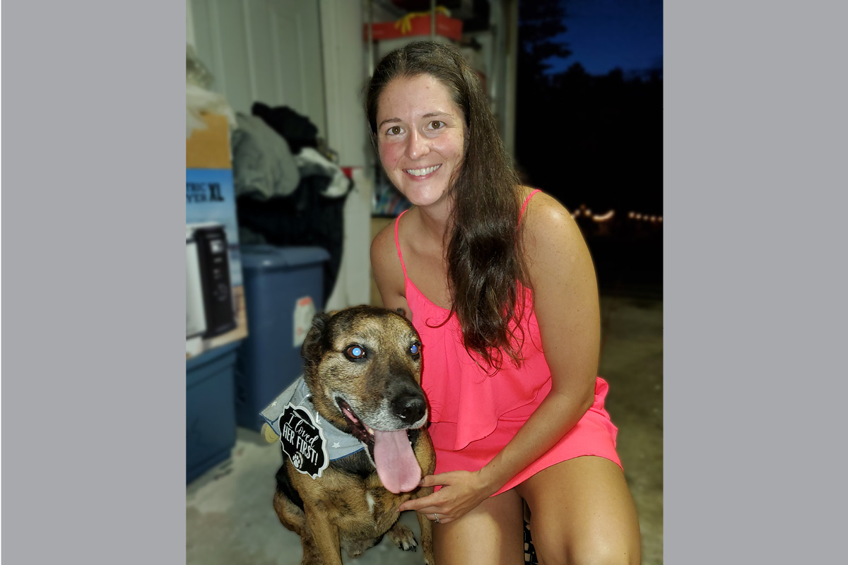 Smiling woman in a pink sleeveless dress sits beside a brown dog with its tongue out, wearing a vest and a badge.
