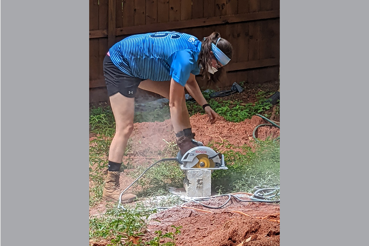 Person wearing a blue striped shirt and safety gear uses a Bosch circular saw to cut a concrete block outdoors.