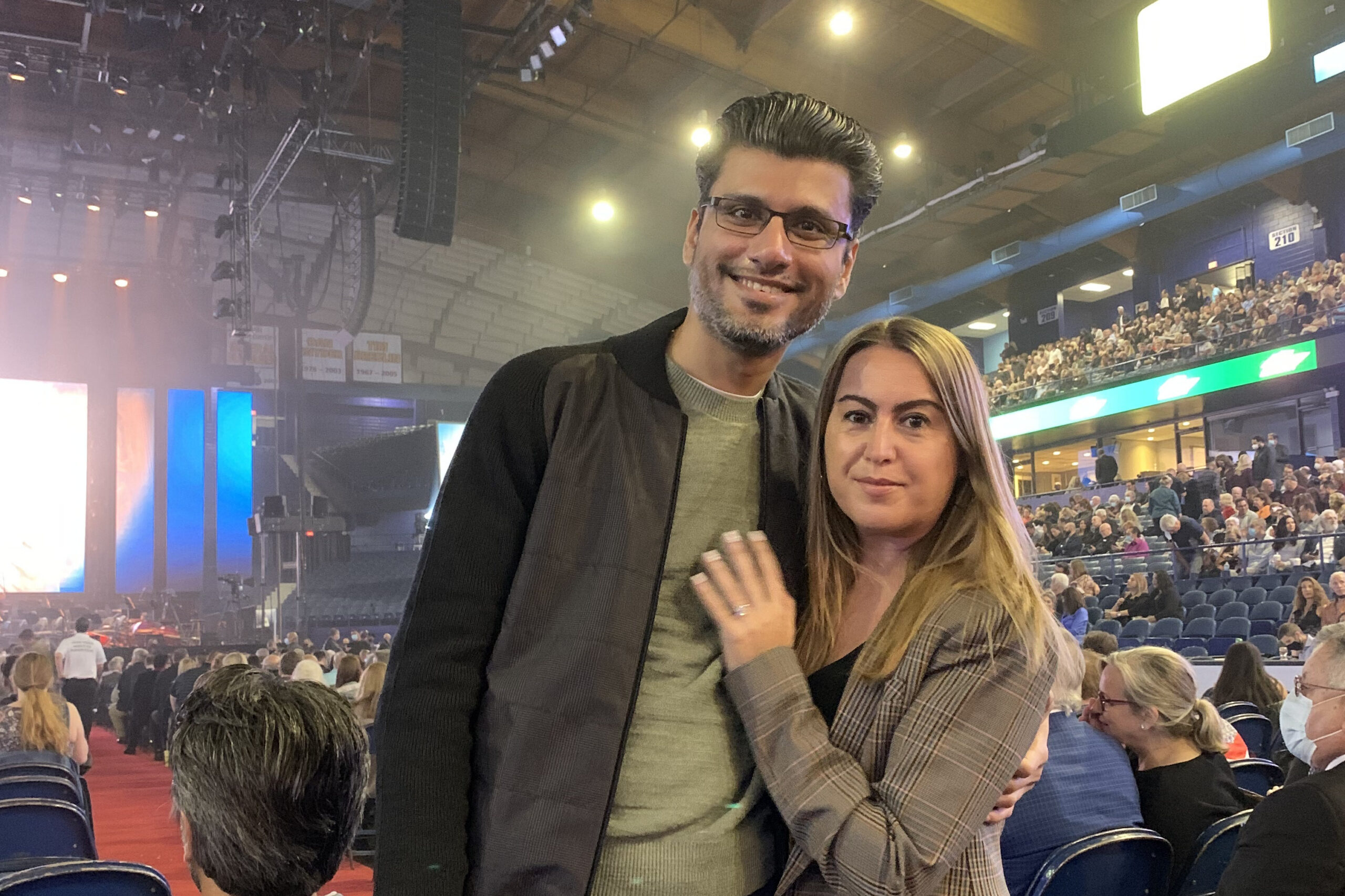 Two people pose for a photo in a crowded indoor arena with stage lighting and a large audience in the background.