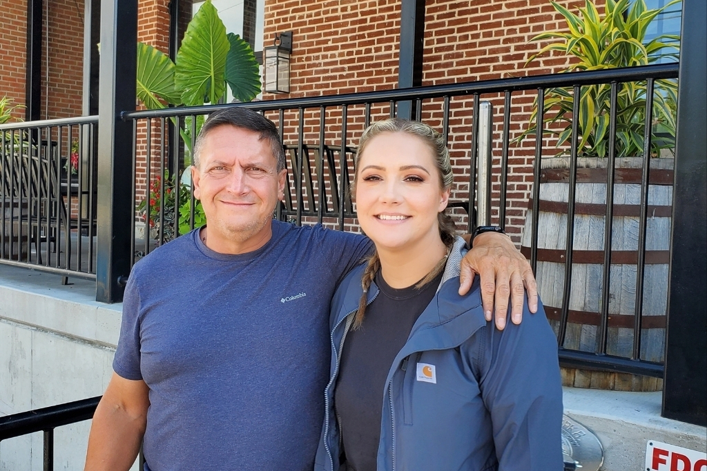 Smiling man and woman stand arm in arm outside a brick building with a black railing behind them.