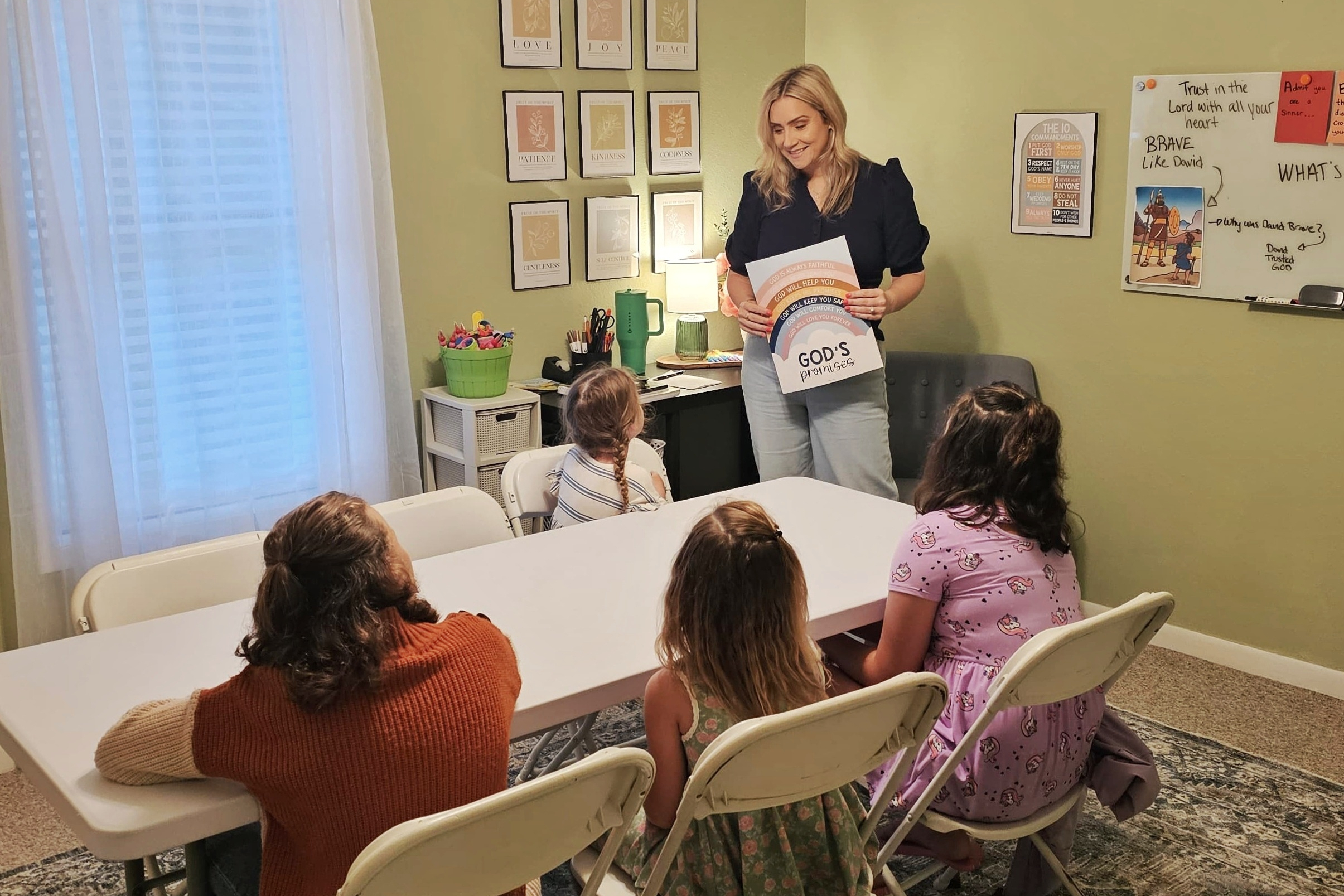 Teacher presenting a circular 'God's Promises' handout to attentive children in a classroom setting with wall posters nearby.