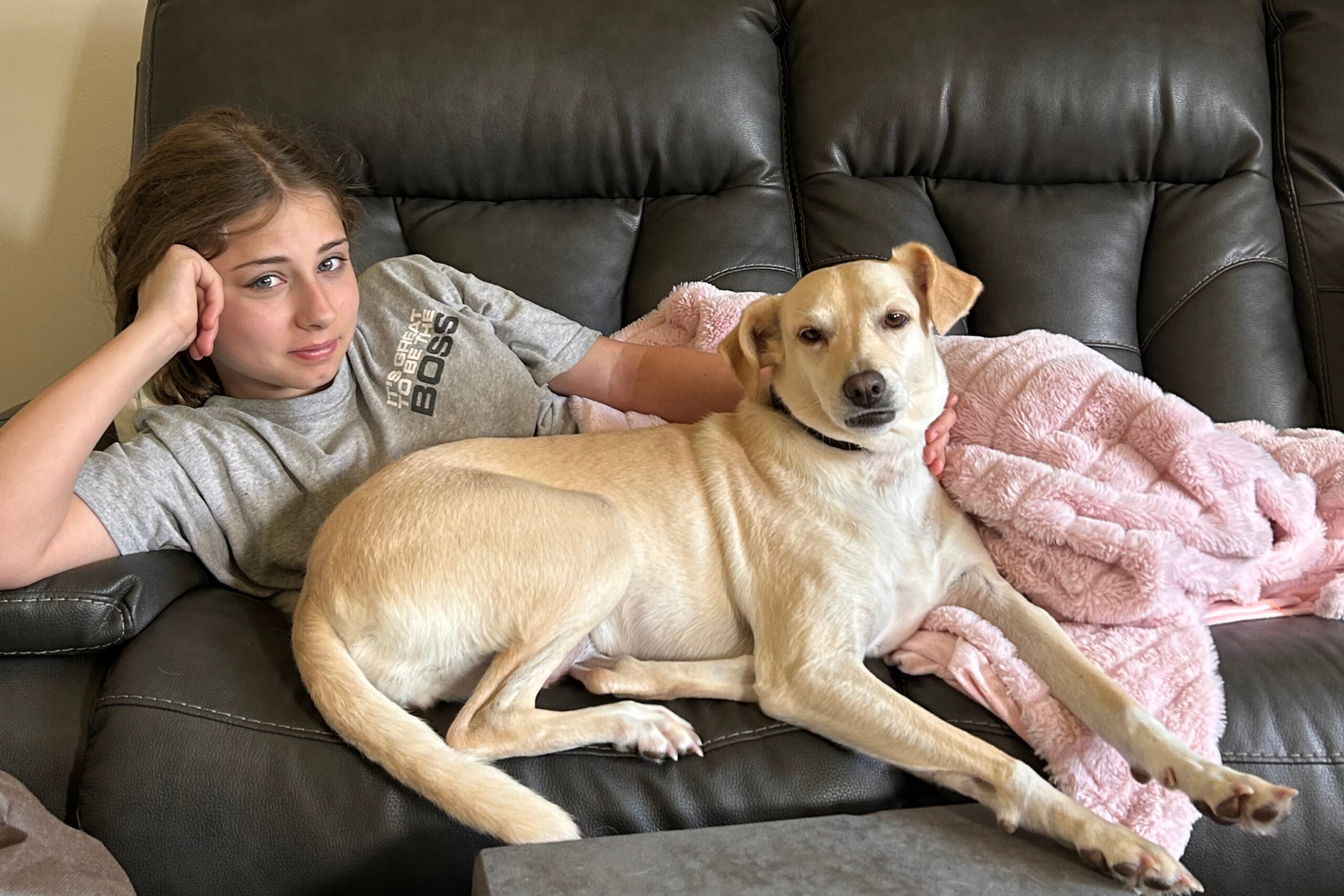 Girl lounging on a black leather couch with a light-colored dog resting on a pink blanket beside her.