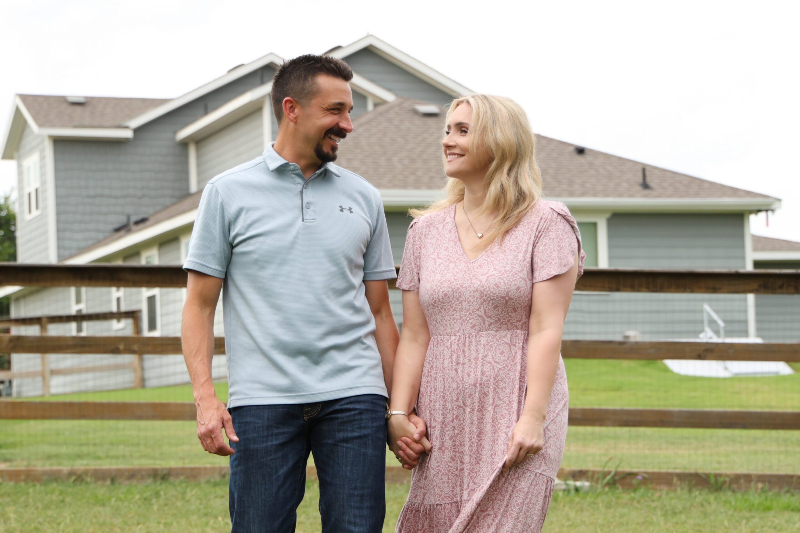 Couple standing in a backyard, holding hands and smiling, with a gray house and wooden fence in the background.