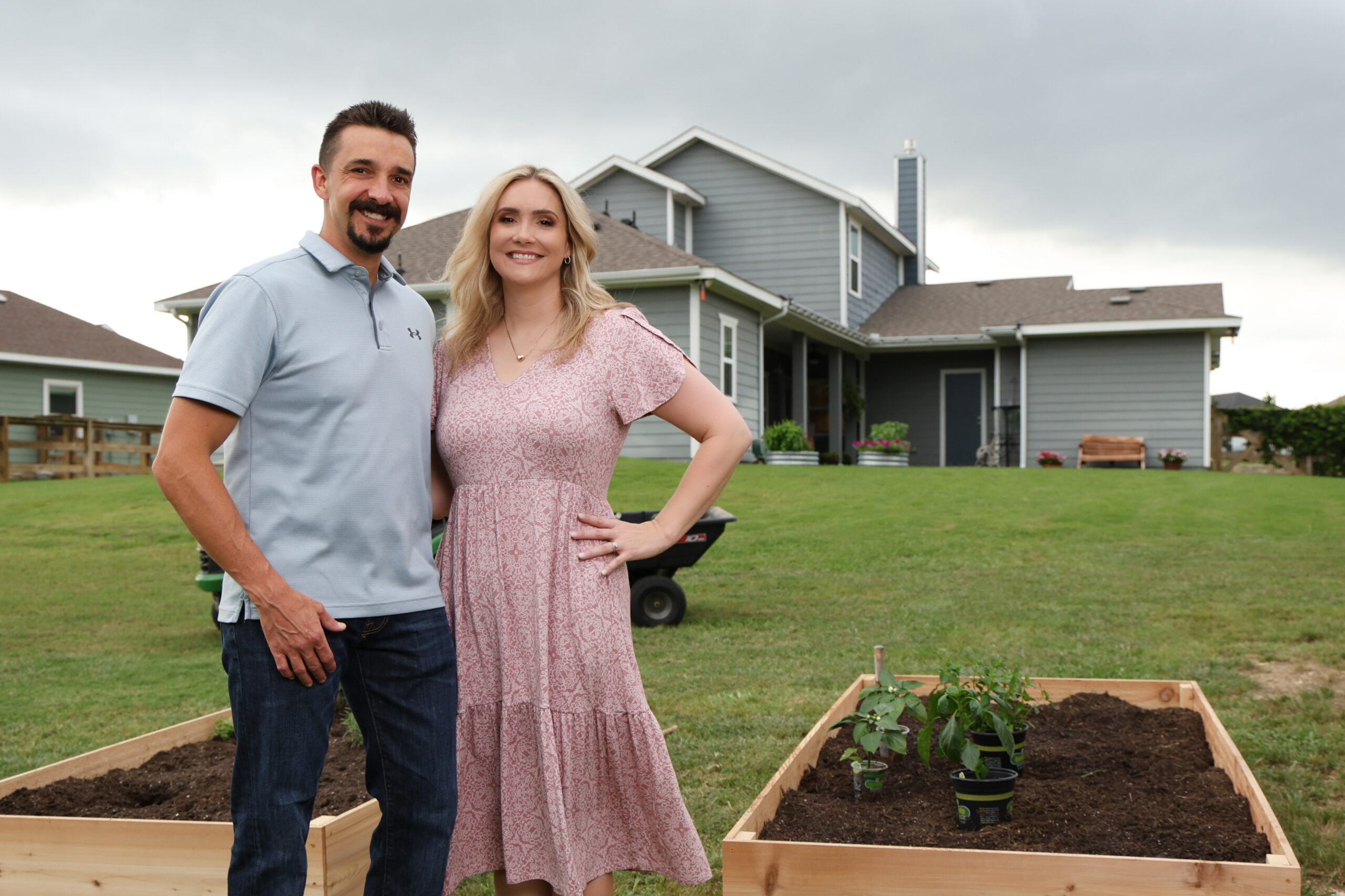 Couple smiling in front of a suburban home with raised garden beds and potted plants in the yard.
