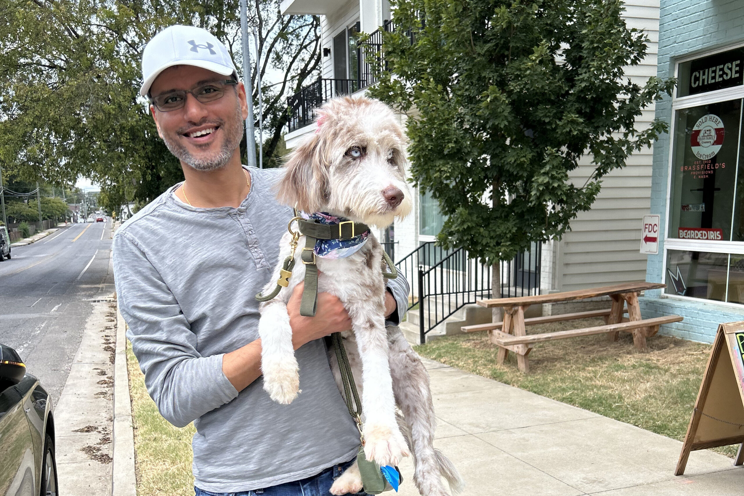 Smiling man in a white cap holding a fluffy light-colored dog on a sidewalk with a cheese shop window in the background.