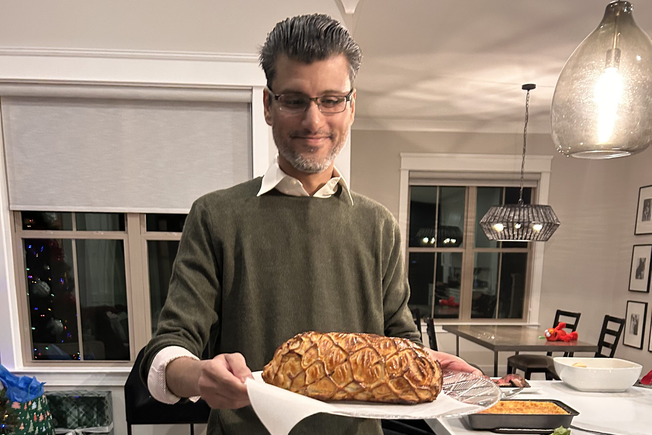 Man in a sweater presents a large braided loaf of bread on a tray in a bright home kitchen.