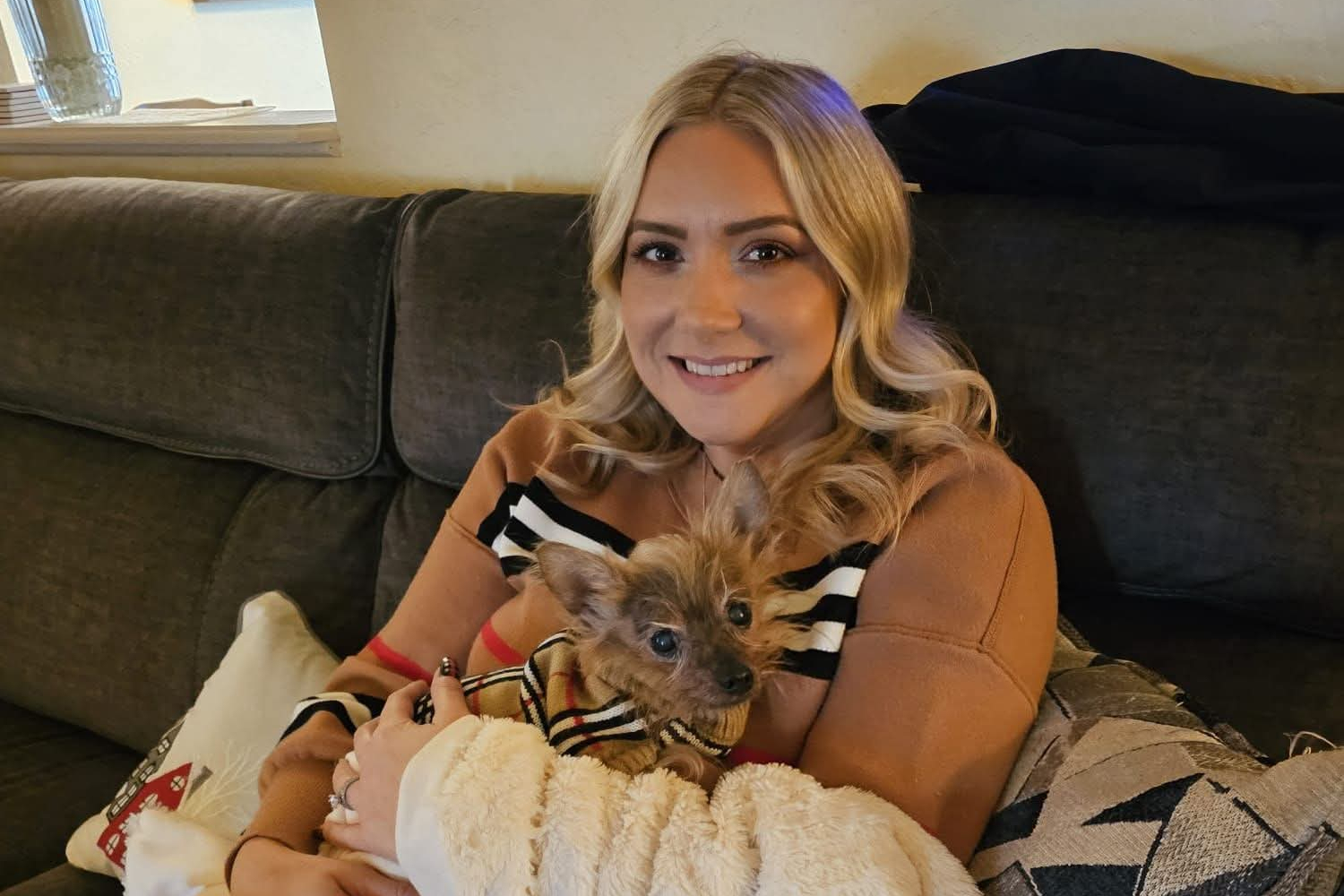 Woman with blonde hair sitting on a sofa, cuddling a small fluffy dog wearing a striped sweater.