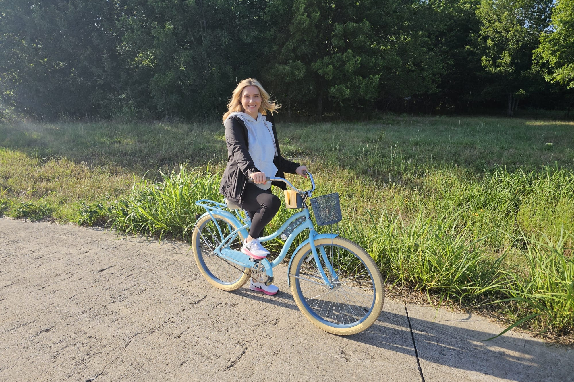 Woman on a pastel blue bicycle riding along a sunny path with tall grass and trees, smiling at the camera on a mild day.