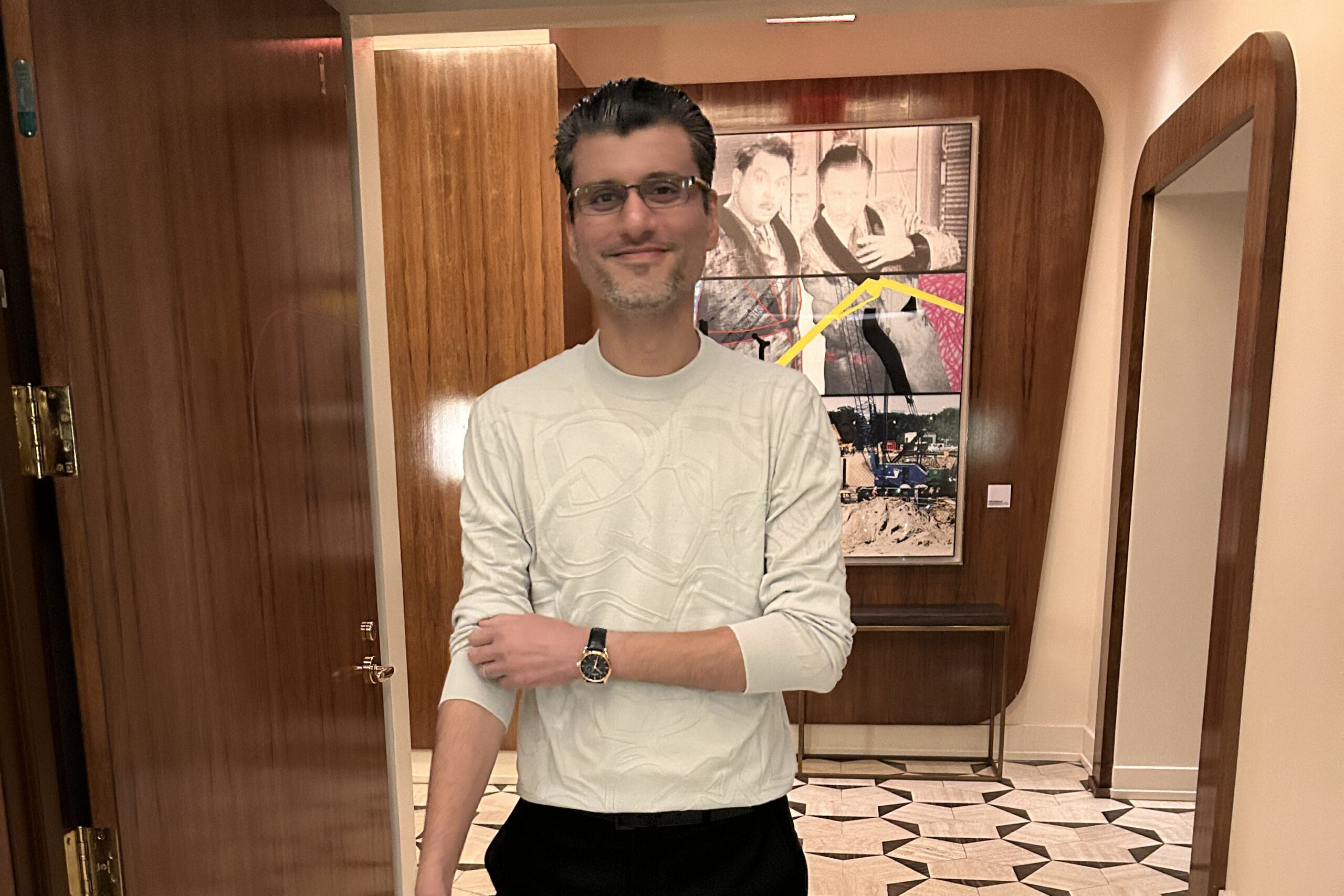 Man in a light gray sweatshirt with glasses stands in a doorway with arms crossed, a poster artwork on the wall behind him.