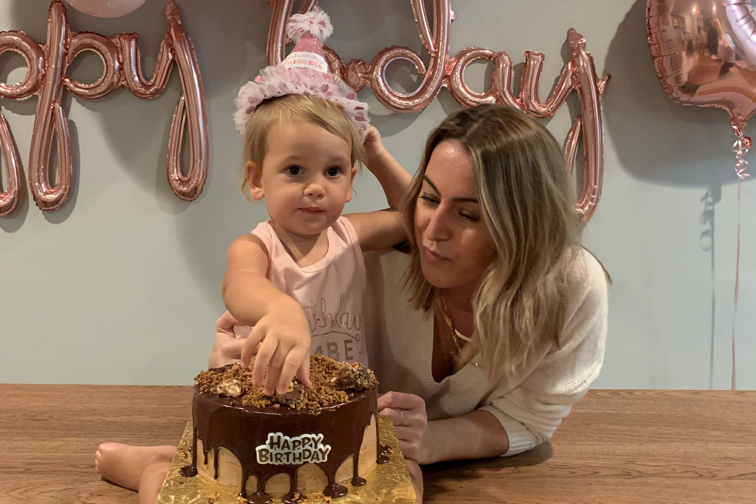 A young girl and a woman cut and touch a chocolate birthday cake at a table, with pink birthday balloons in the background.