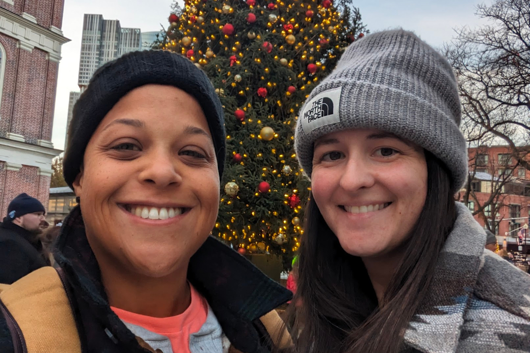 Two smiling women take a selfie in front of a large, decorated Christmas tree in an urban plaza at dusk.