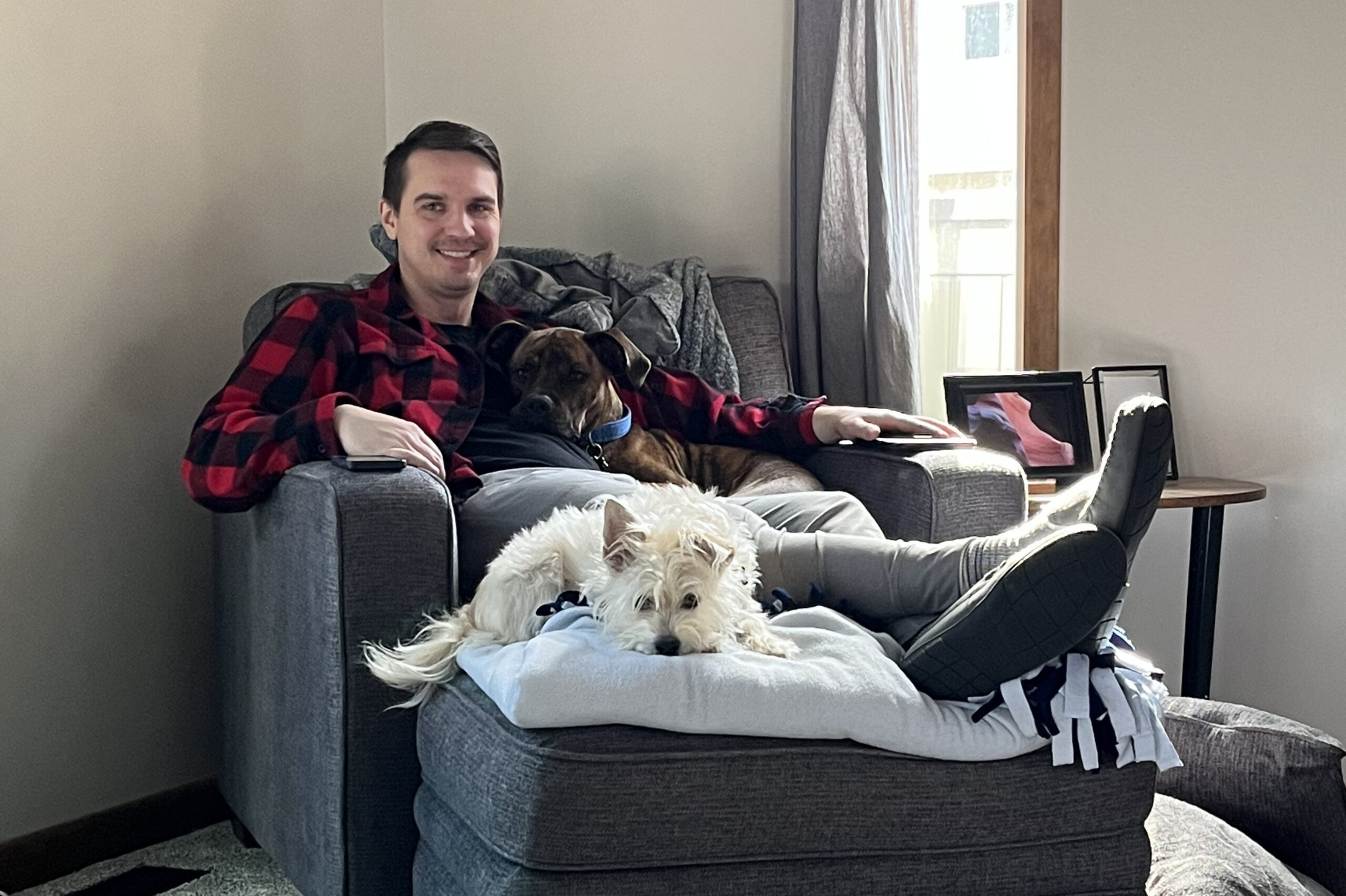 Man in red plaid shirt relaxing on a gray sofa with a small white fluffy dog on his lap in a living room. A window with curtains and framed photos are in the background.