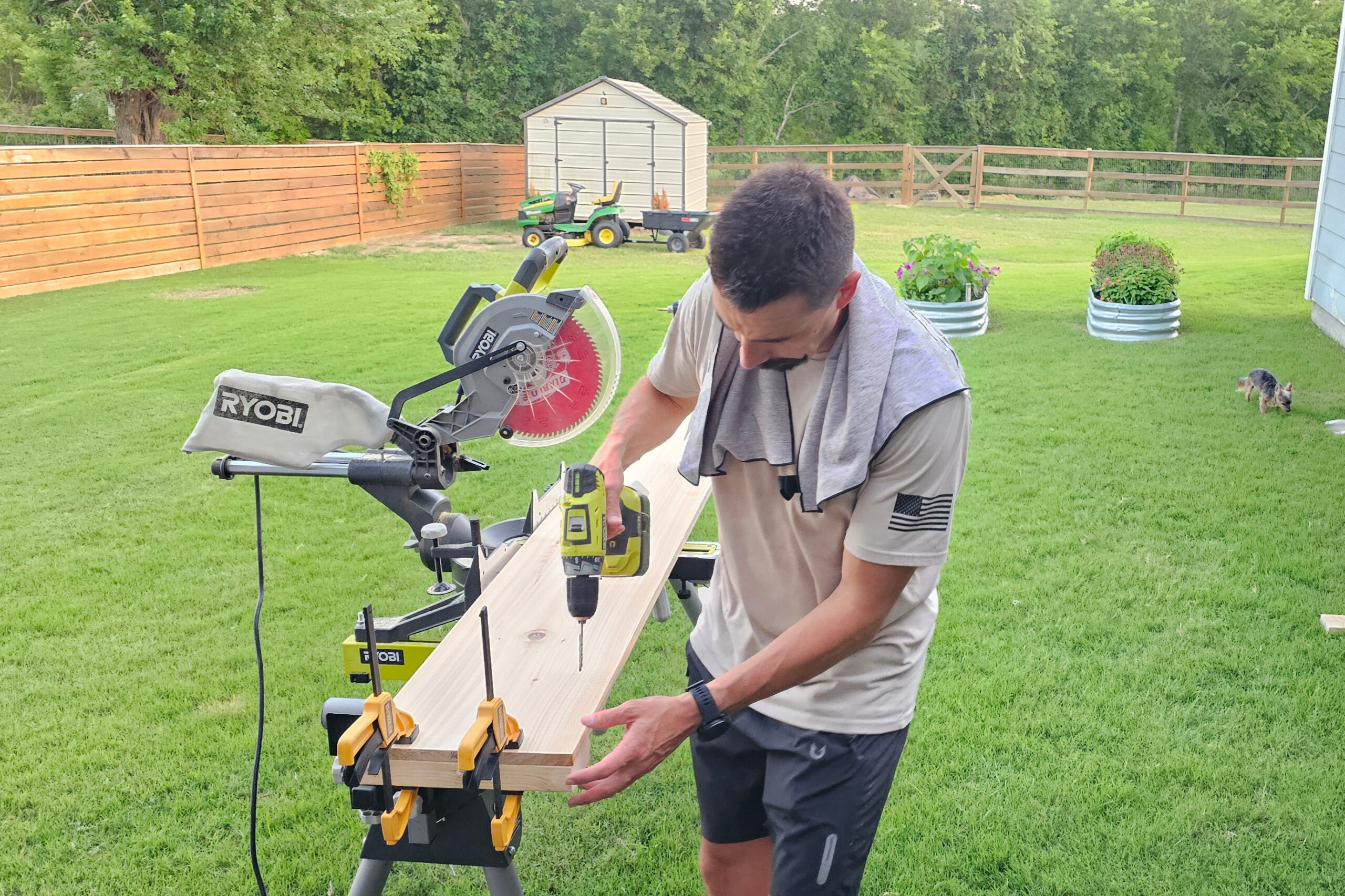 Man in a light shirt and shorts cuts a long wooden board on a miter saw in a backyard, with a shed and green lawn in the background.