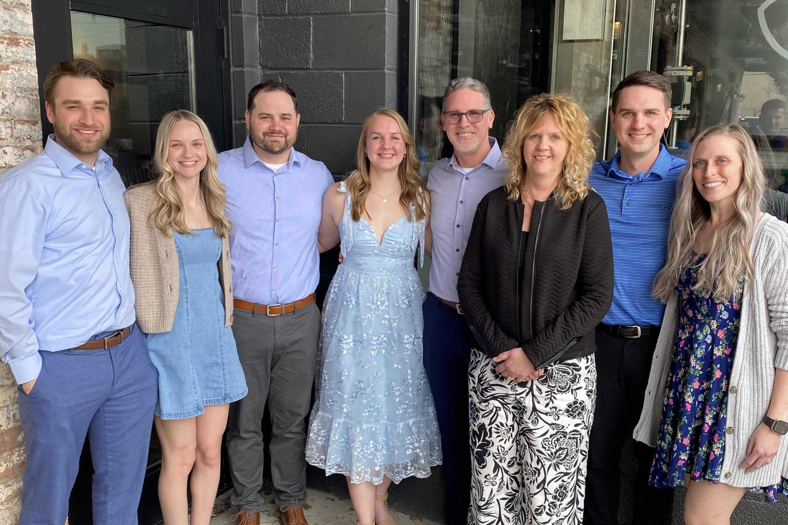 Eight adults standing close together outdoors in front of a dark storefront, smiling for a group photo, wearing casual to semi-formal attire.