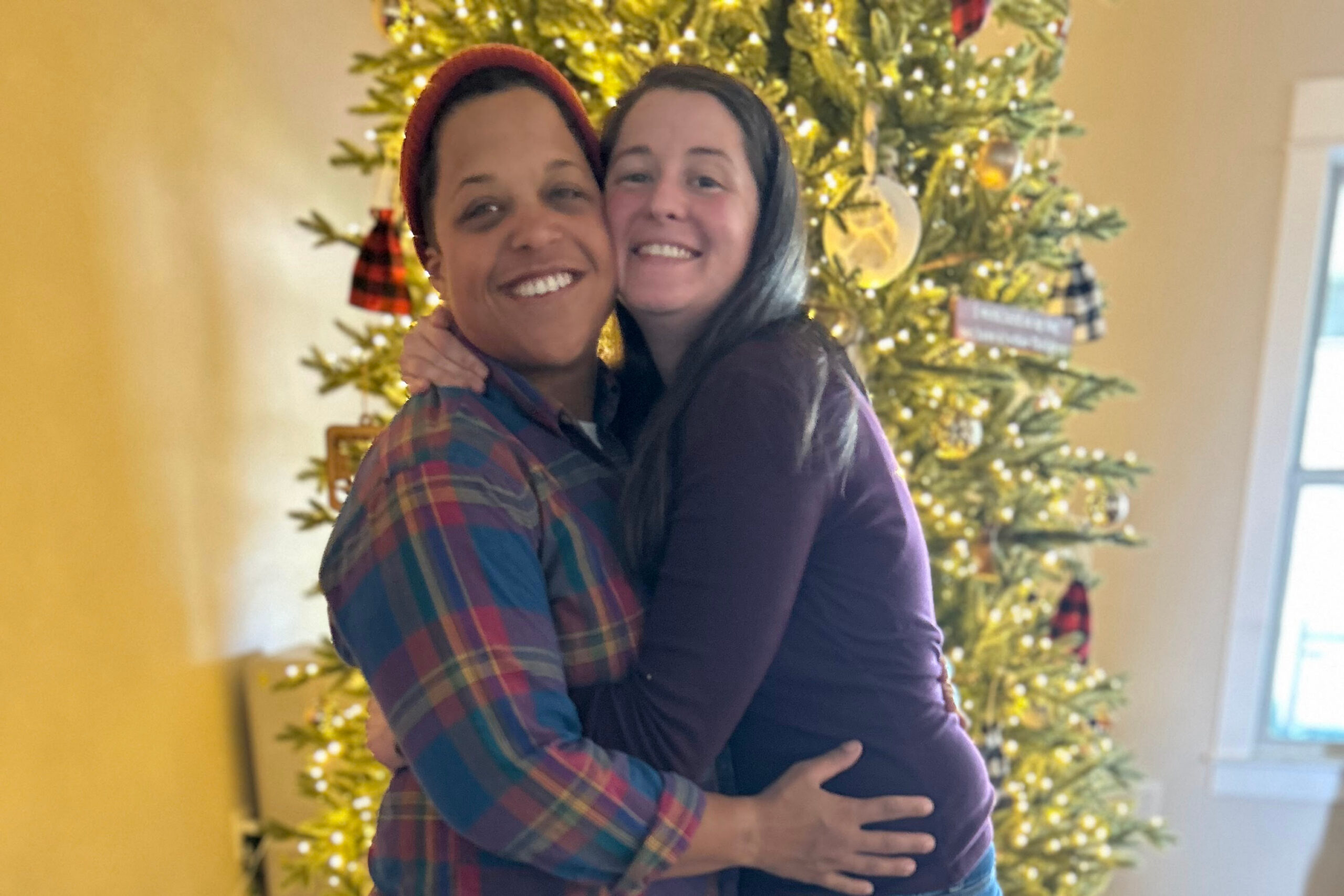 Two people hugging and smiling in front of a lit Christmas tree indoors, festive scene behind them.