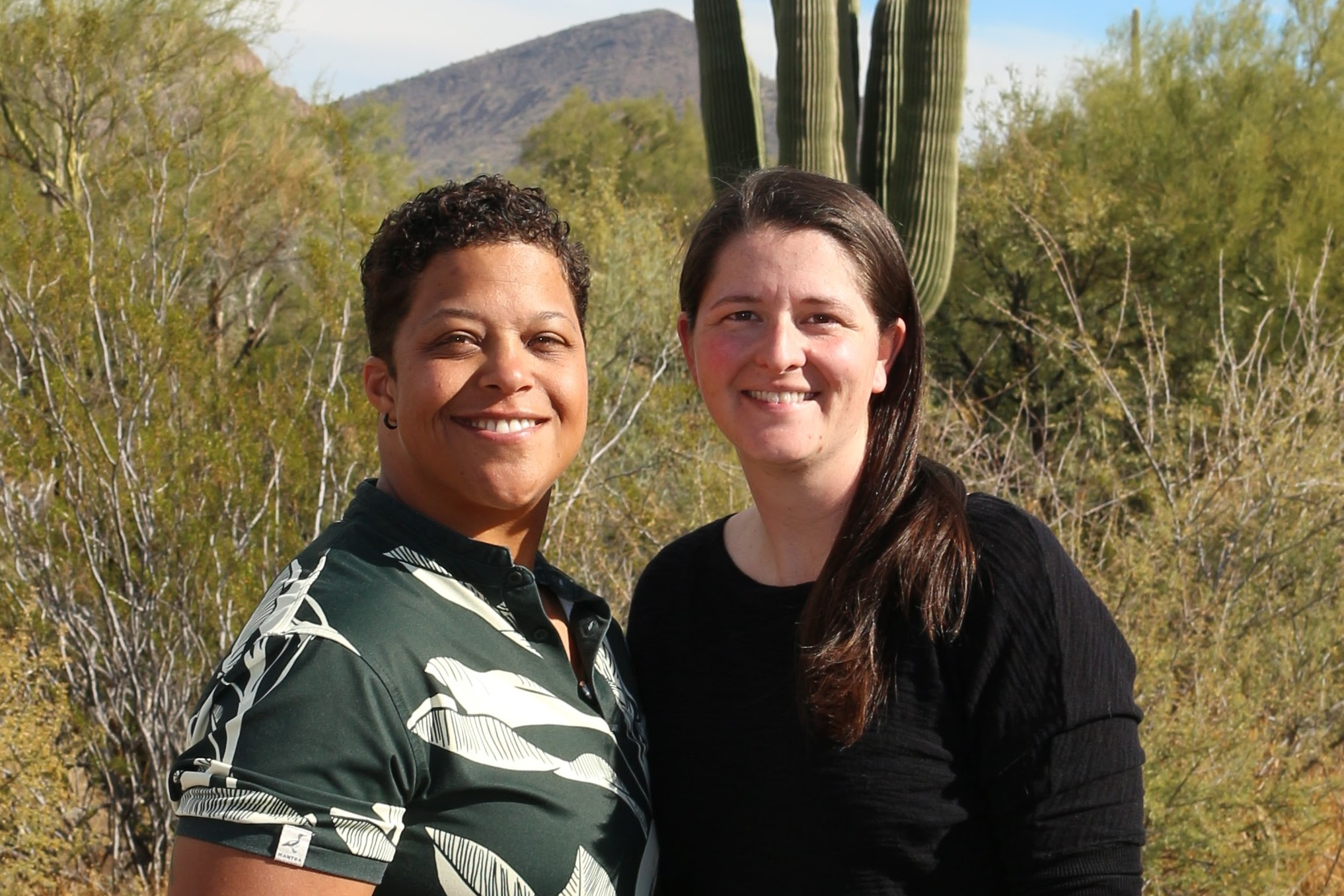 Two women smiling and posing together outdoors in a desert with tall cacti and hills in the background, daytime sun.