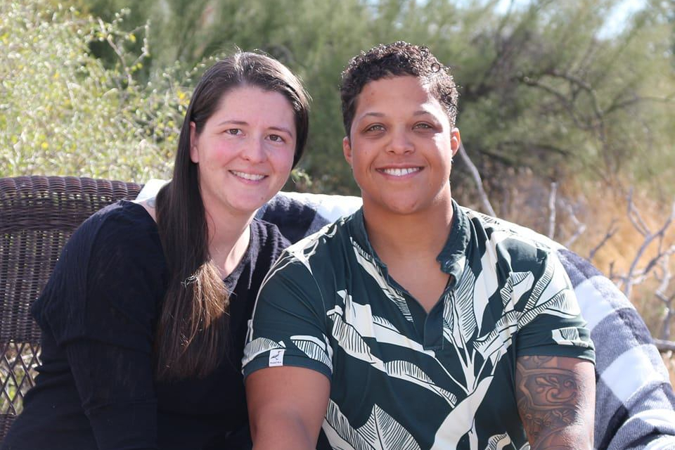 Two friends sit close together on a wicker bench outdoors, smiling at the camera with greenery in the background.