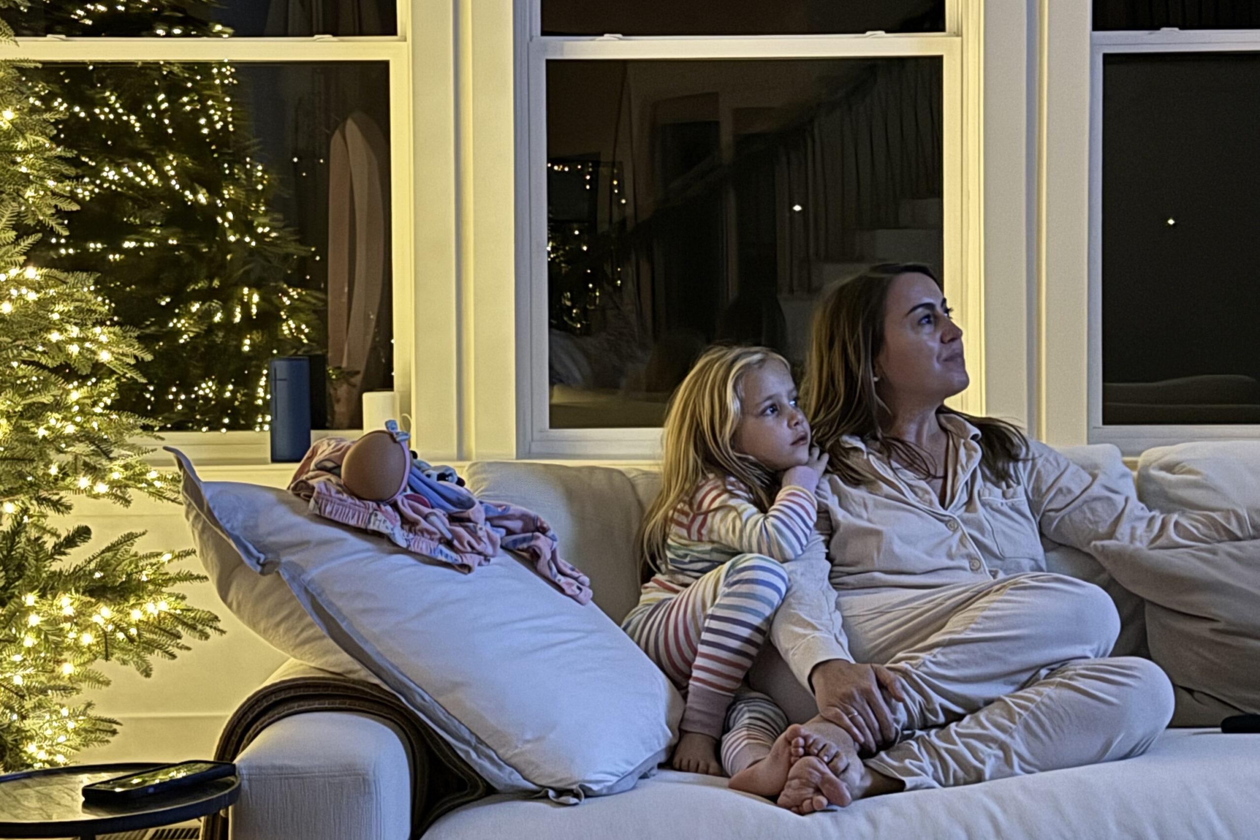 Mother and young daughter cuddled on a beige sofa near a lit Christmas tree with twinkling lights.