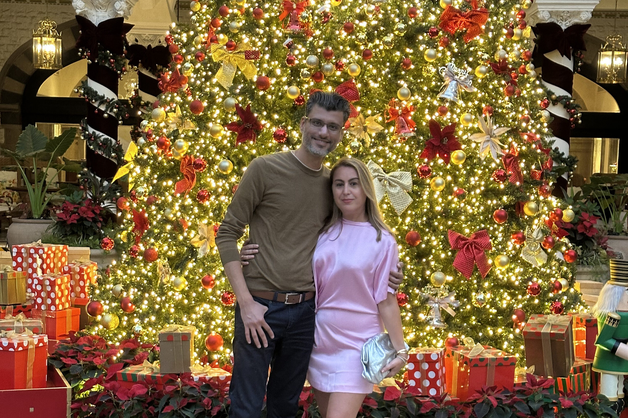 Couple posing in front of a large, brightly lit Christmas tree with gold and red ornaments and wrapped presents around the base in a festive indoor setting.