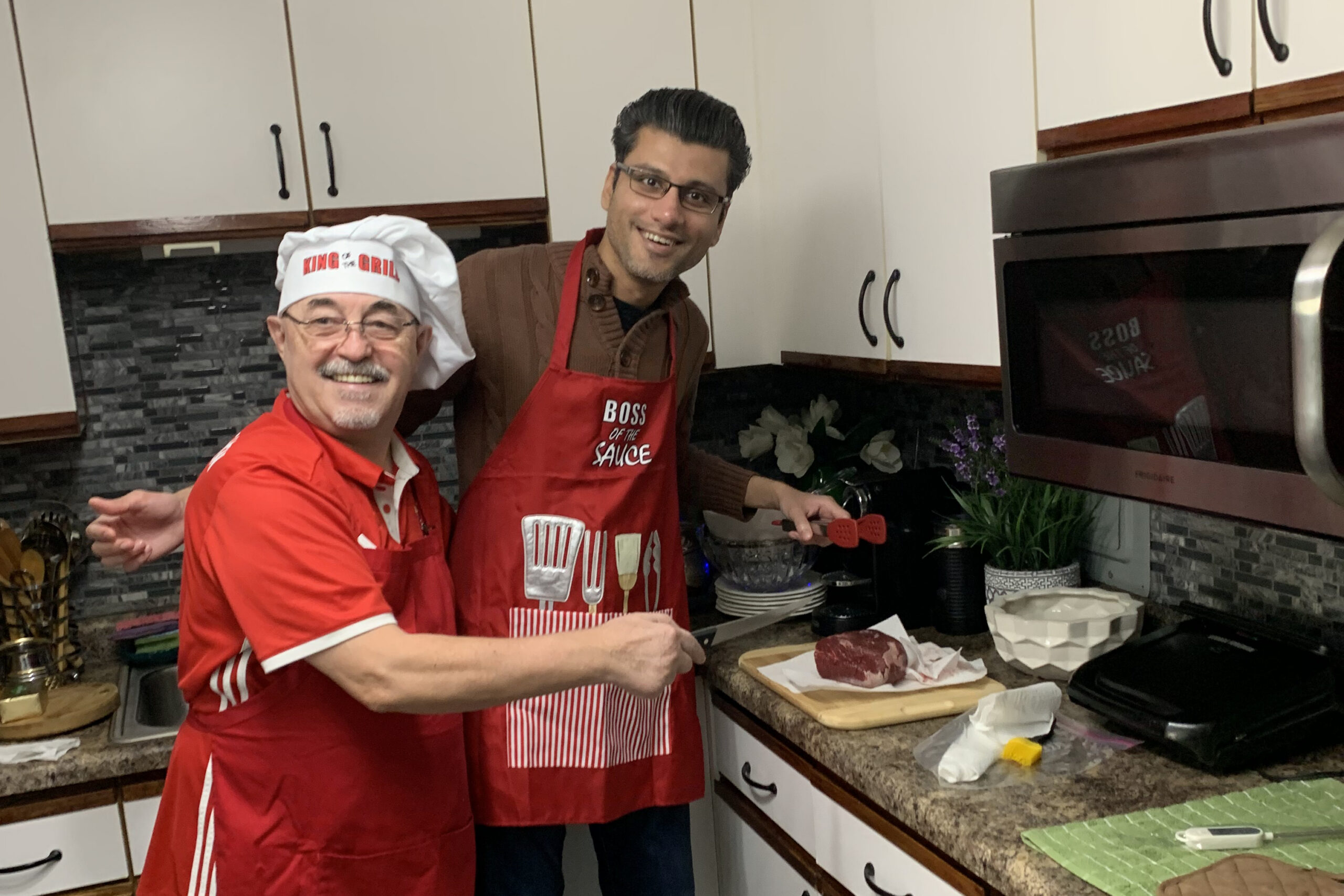 Two men in red aprons grilling in a home kitchen, one wearing a chef’s hat that reads 'King of the Grill'.