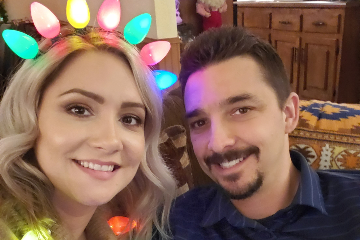 Smiling couple taking a selfie; woman wears a multicolored LED light headband indoors.