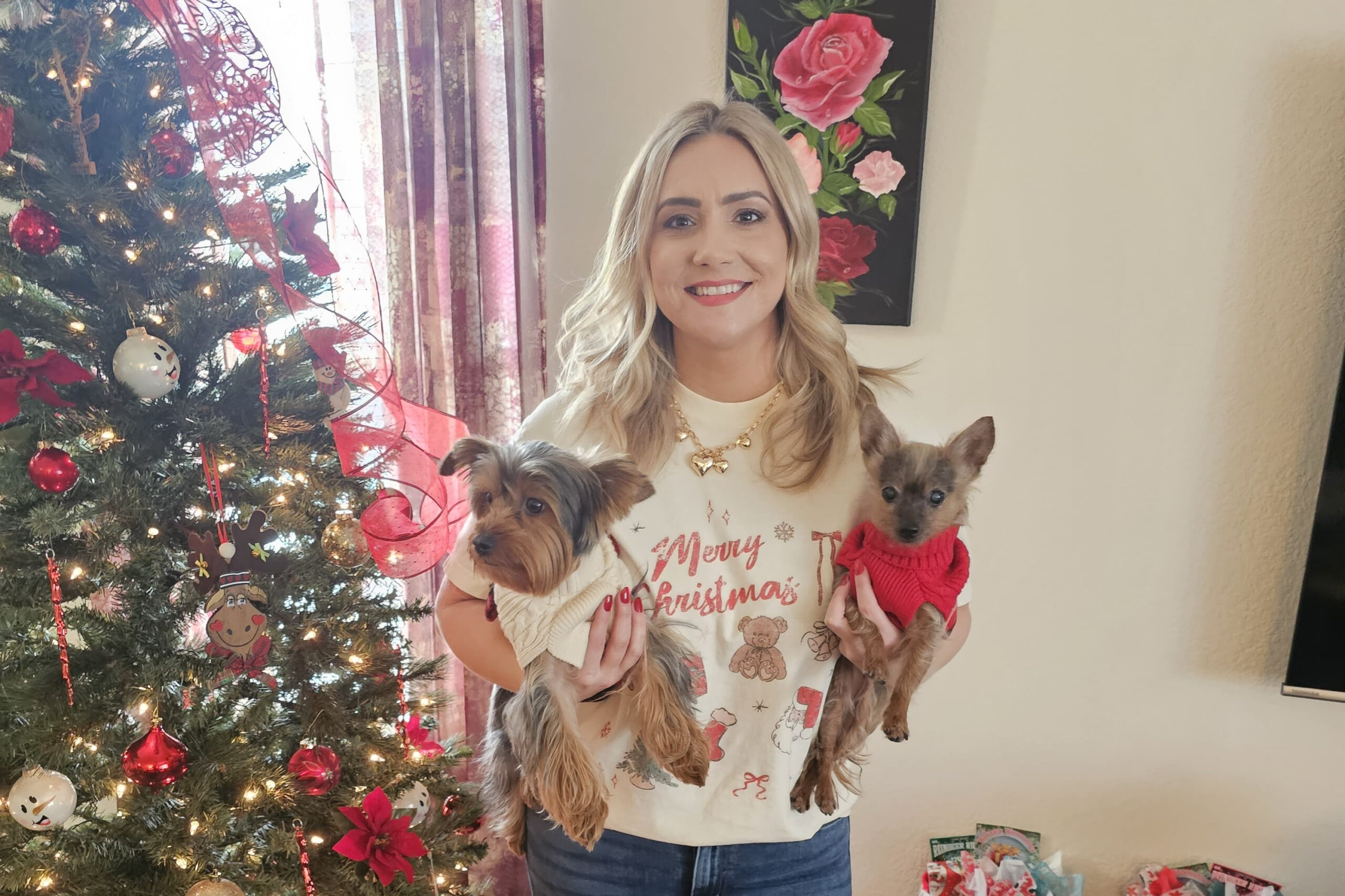 Woman smiling while holding two small dogs in front of a Christmas tree with lights and red decorations.
