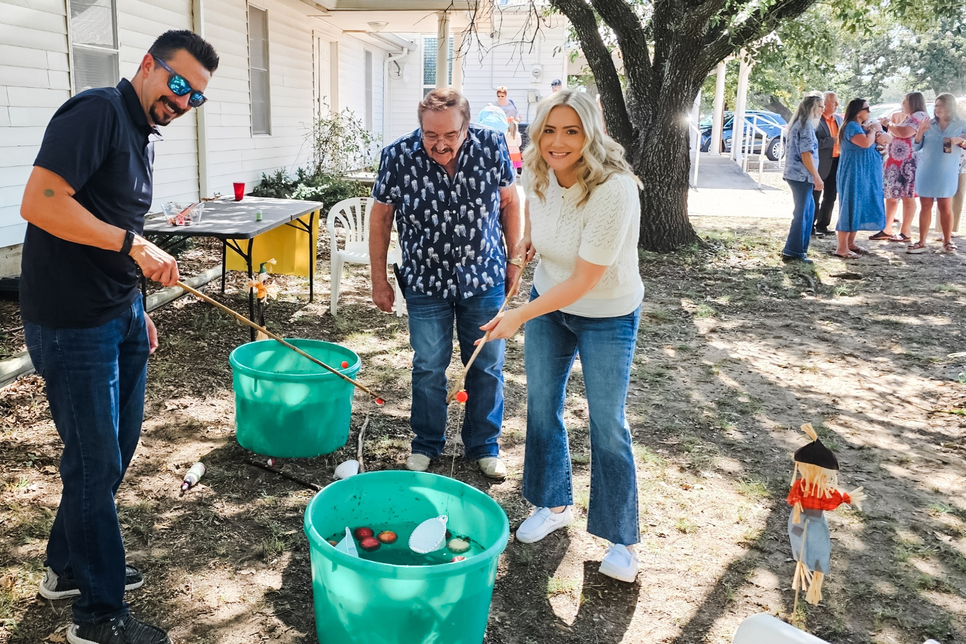 Three people participate in a yard party game with spoons and green buckets while others chat in the background under a tree outside a house.