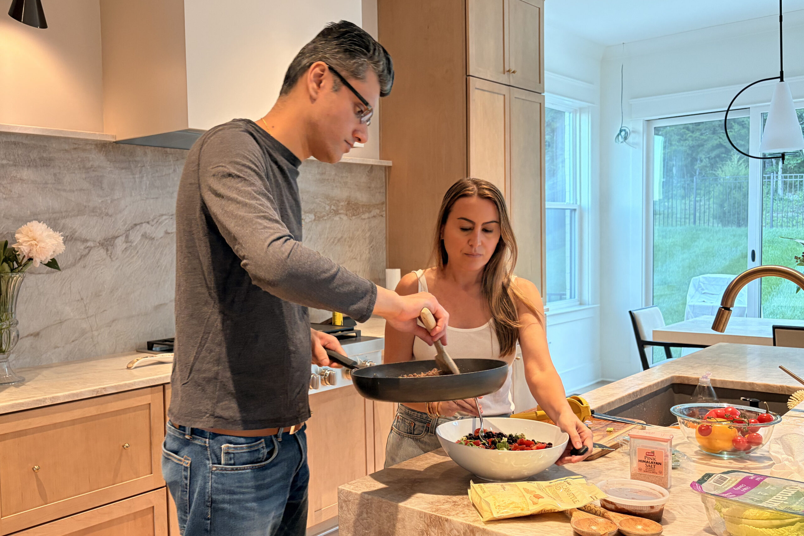 Man and woman cooking together in a bright modern kitchen, stirring ingredients in a large pan on a marble island.