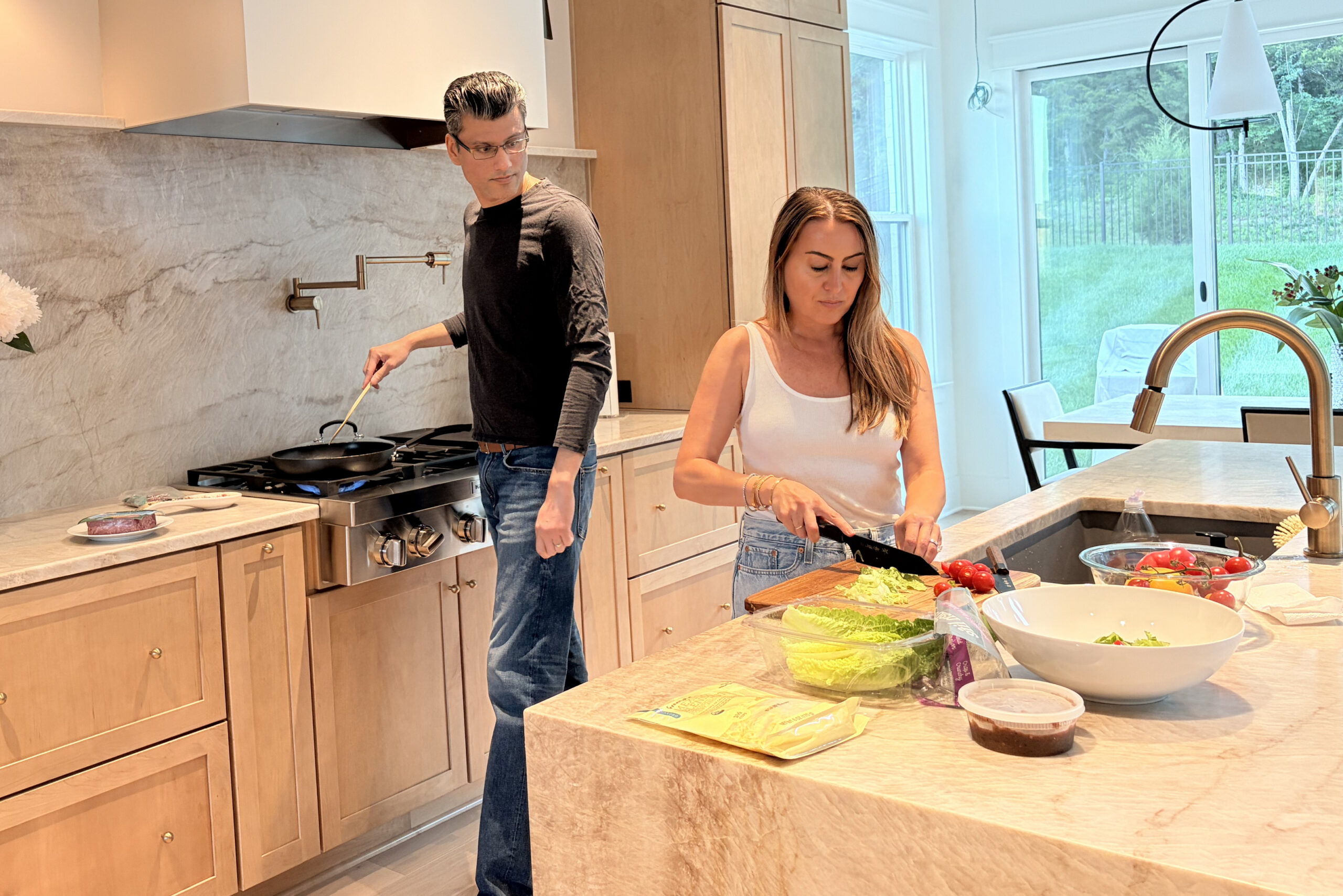 Man cooking at a stove while a woman chops lettuce and tomato slices at a marble kitchen island in a bright, modern kitchen.
