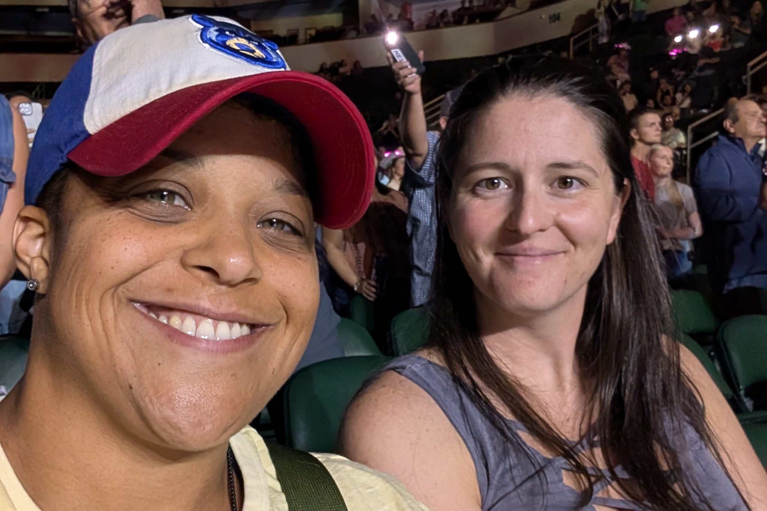 Two women taking a selfie at a crowded arena, smiling at the camera, with one wearing a white and blue cap and the other with dark hair and a green top.