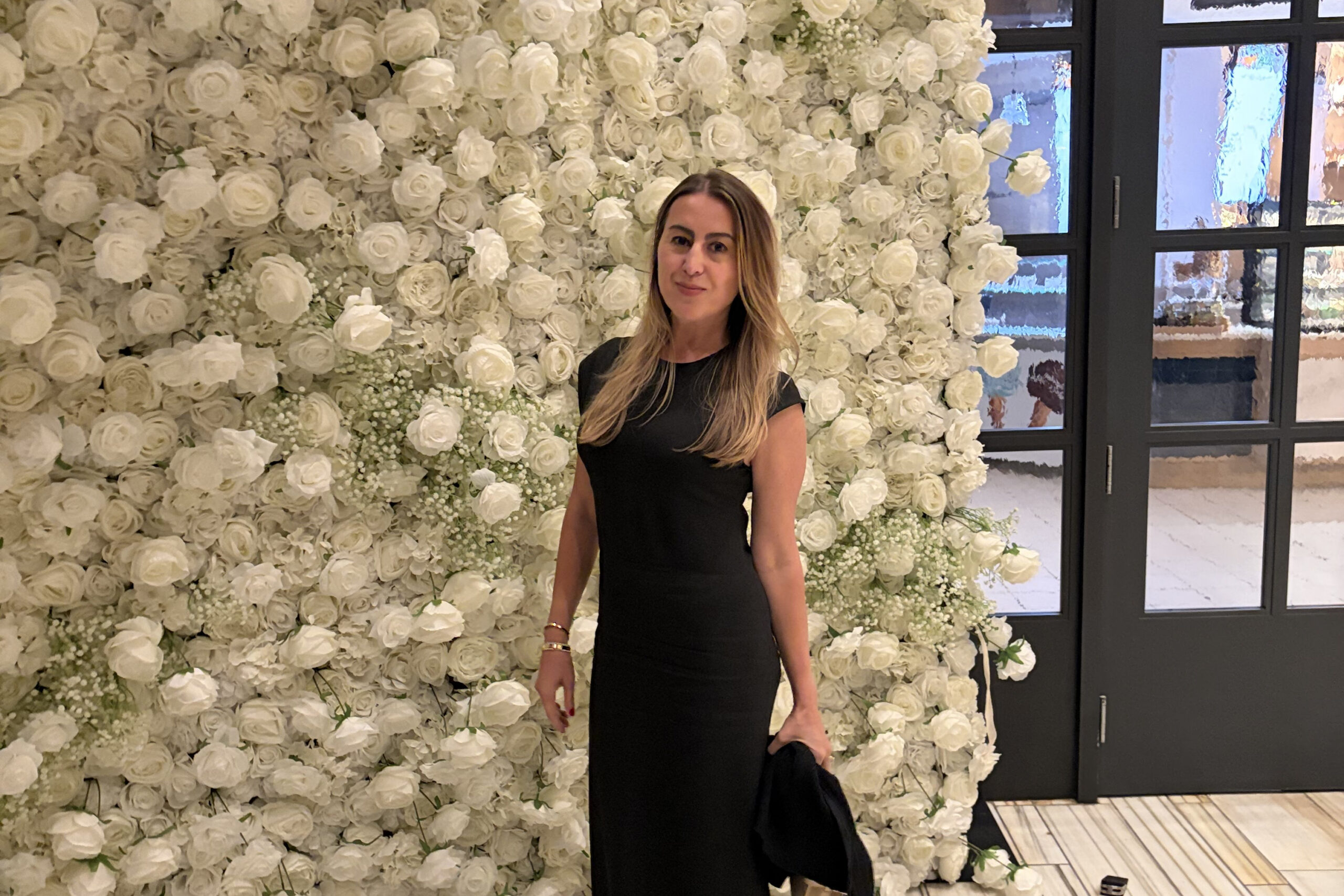 Woman in a black dress posing in front of a dense wall of white roses at an event, with glass doors to the right.