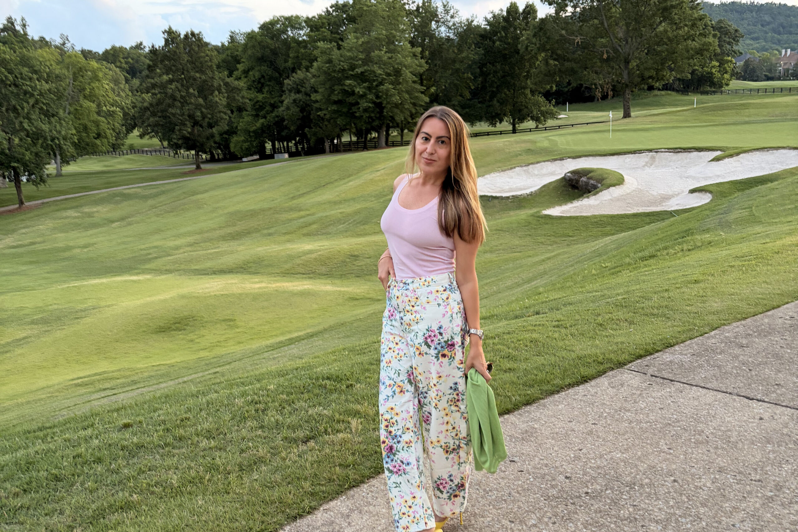 Woman wearing a pink sleeveless top and floral pants posing on a golf course path with a sand bunker behind her.