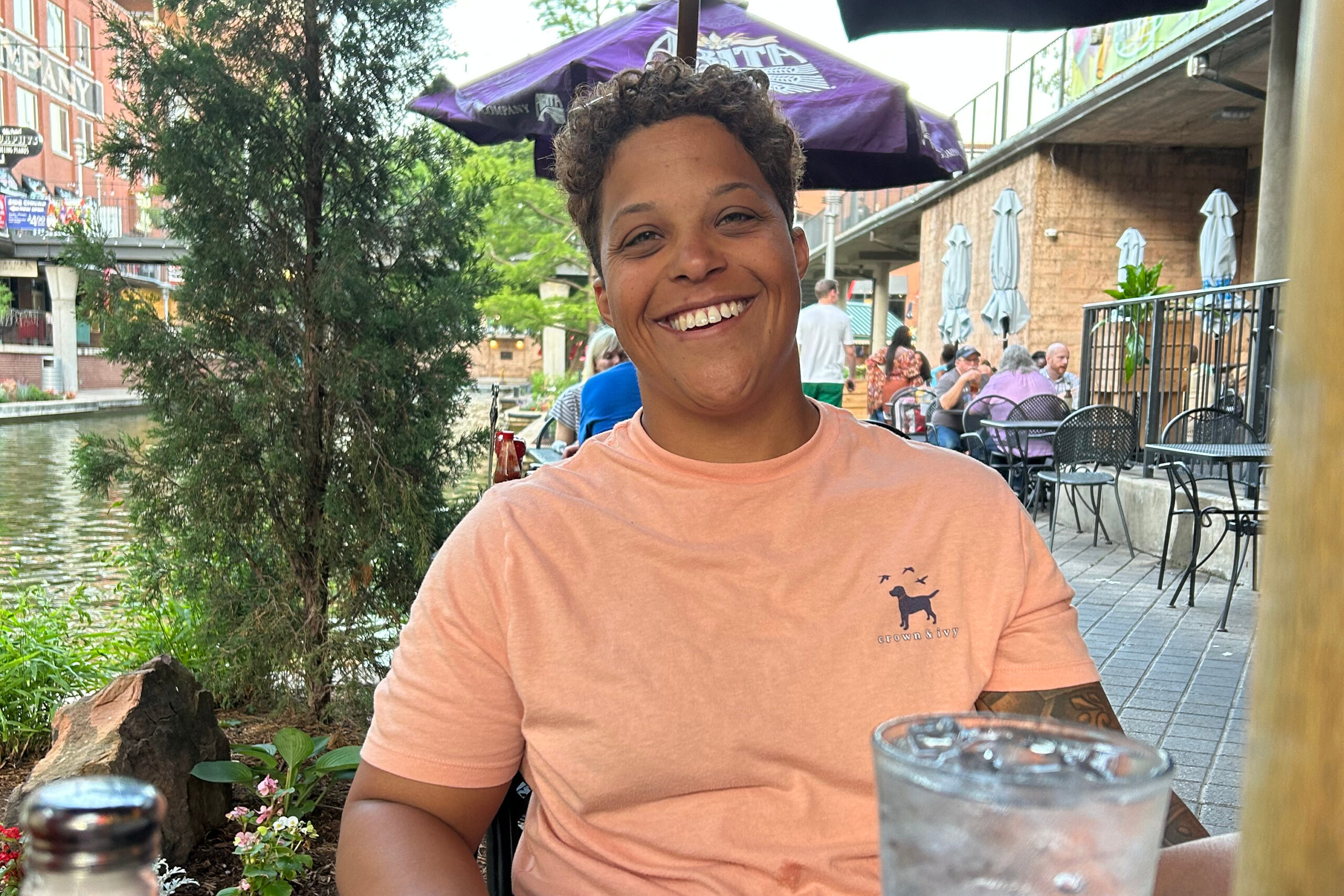 Smiling person in a peach T-shirt sits at an outdoor cafe by a canal, with drinks on the table in the foreground.
