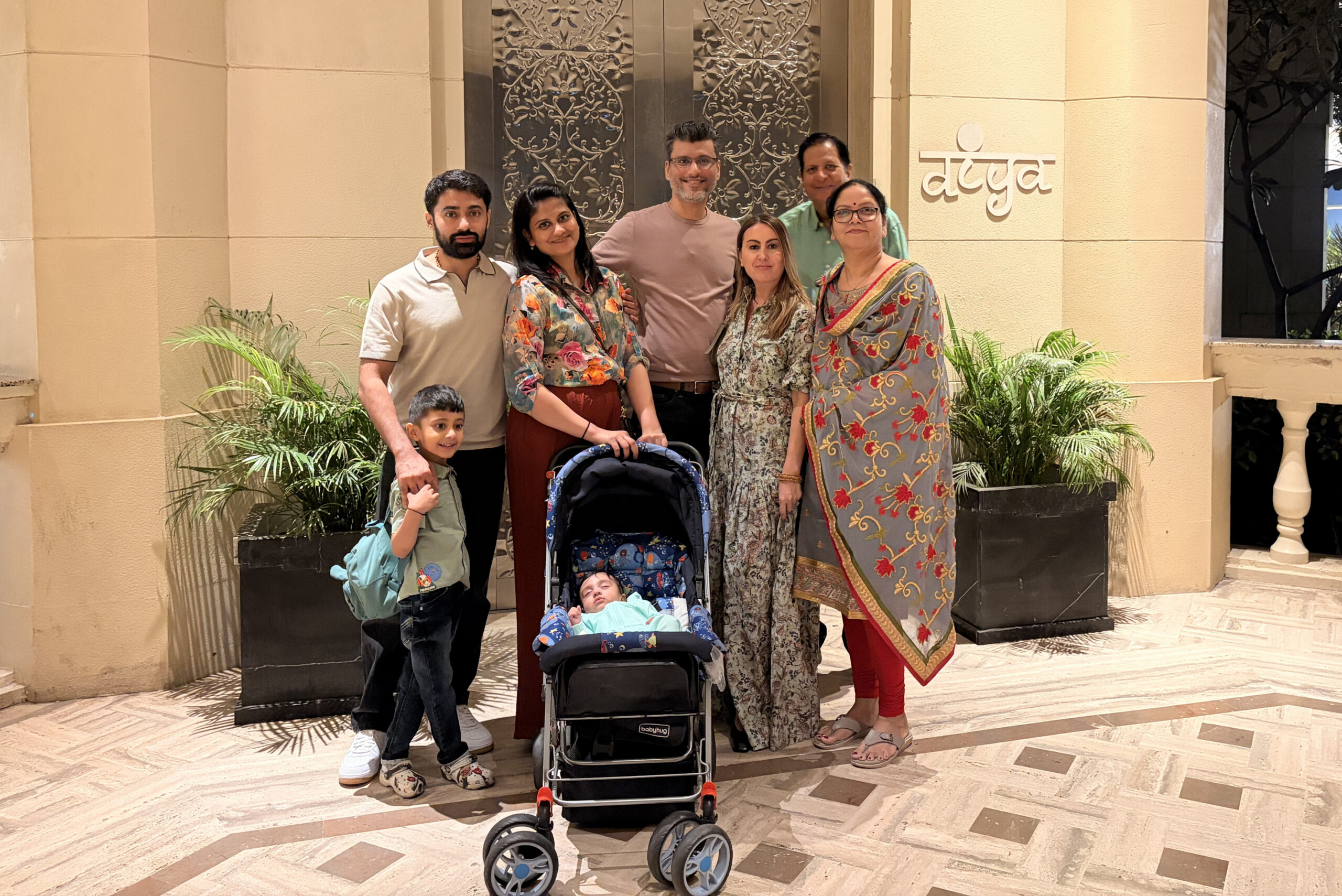 Group of six adults with a baby in a stroller posing in a beige hotel lobby with a decorative door in the background, all smiling.